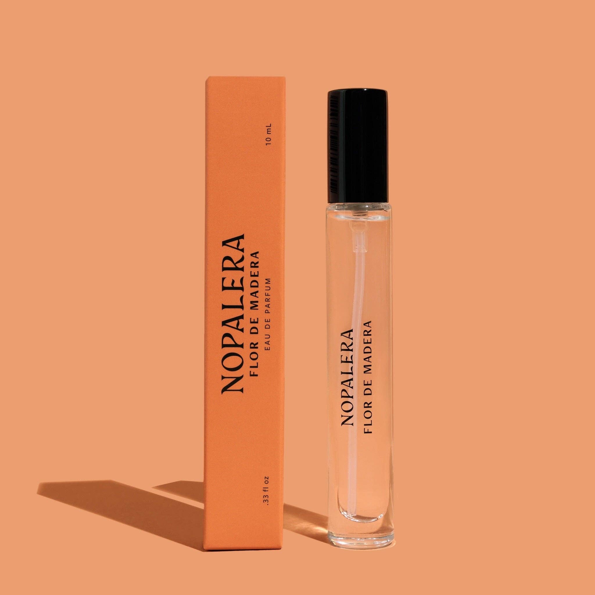 A clear glass 10mL Flor de Madera Eau de Parfum Travel Spray by Nopalera, with a black cap, is displayed beside its matching orange box on an orange background. Both cast soft shadows to the left.