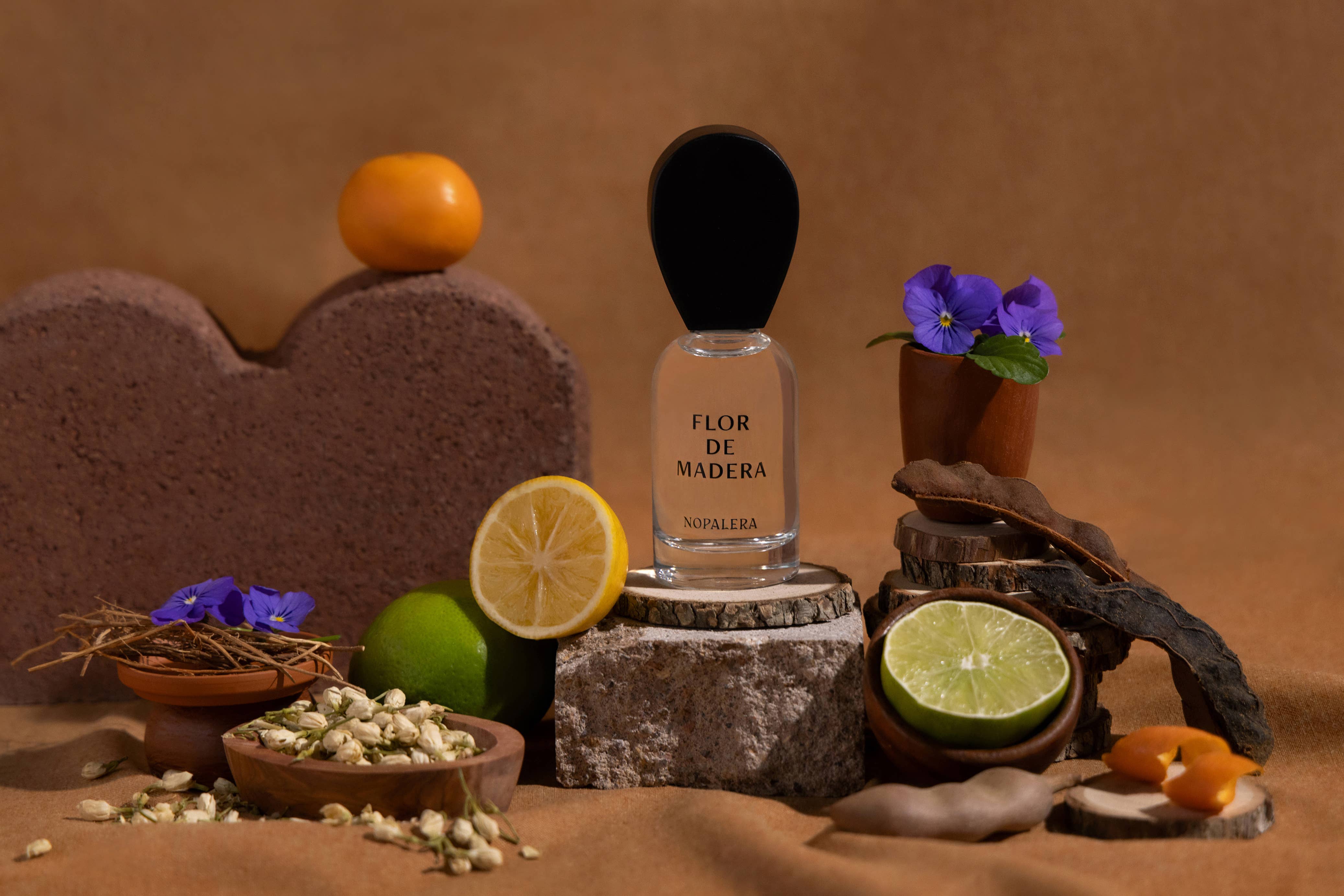 A 10mL Flor de Madera Eau de Parfum Travel Spray by Nopalera rests on a stone, encircled by Mexican citrus—lime, lemon, orange—flowers, dried pods, and botanicals against a brown backdrop.