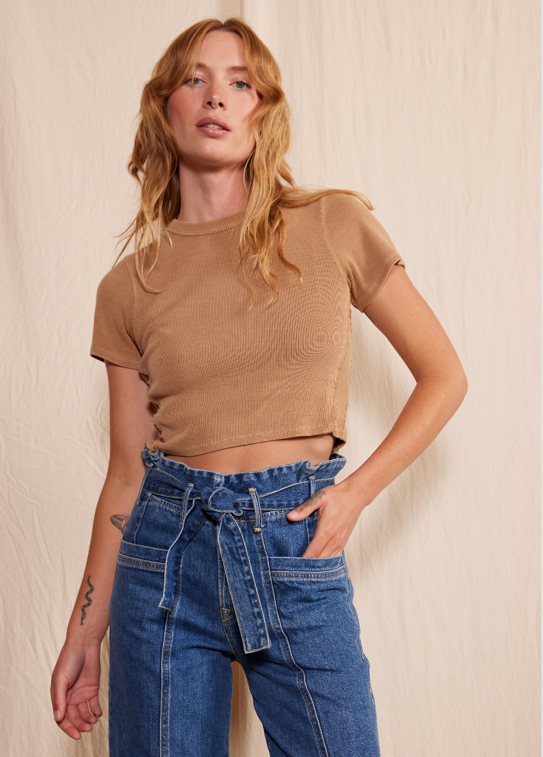 A woman with wavy, light brown hair models Porter Blue Apparels PENNY PAPERBAG WIDE LEG IN LAGUNA WASH jeans and a fitted tan crop top, posing against a beige fabric backdrop with one hand in her pocket.