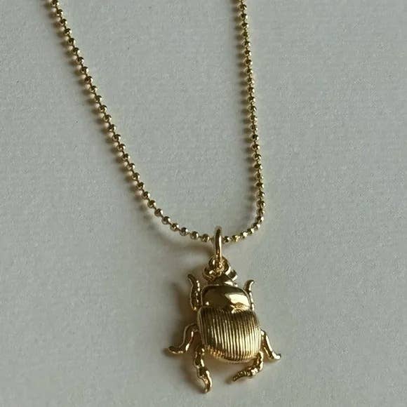 The Scarab Necklace by Tramps+Thieves features a gold-filled scarab beetle pendant on a beaded gold chain, elegantly displayed on a light surface.
