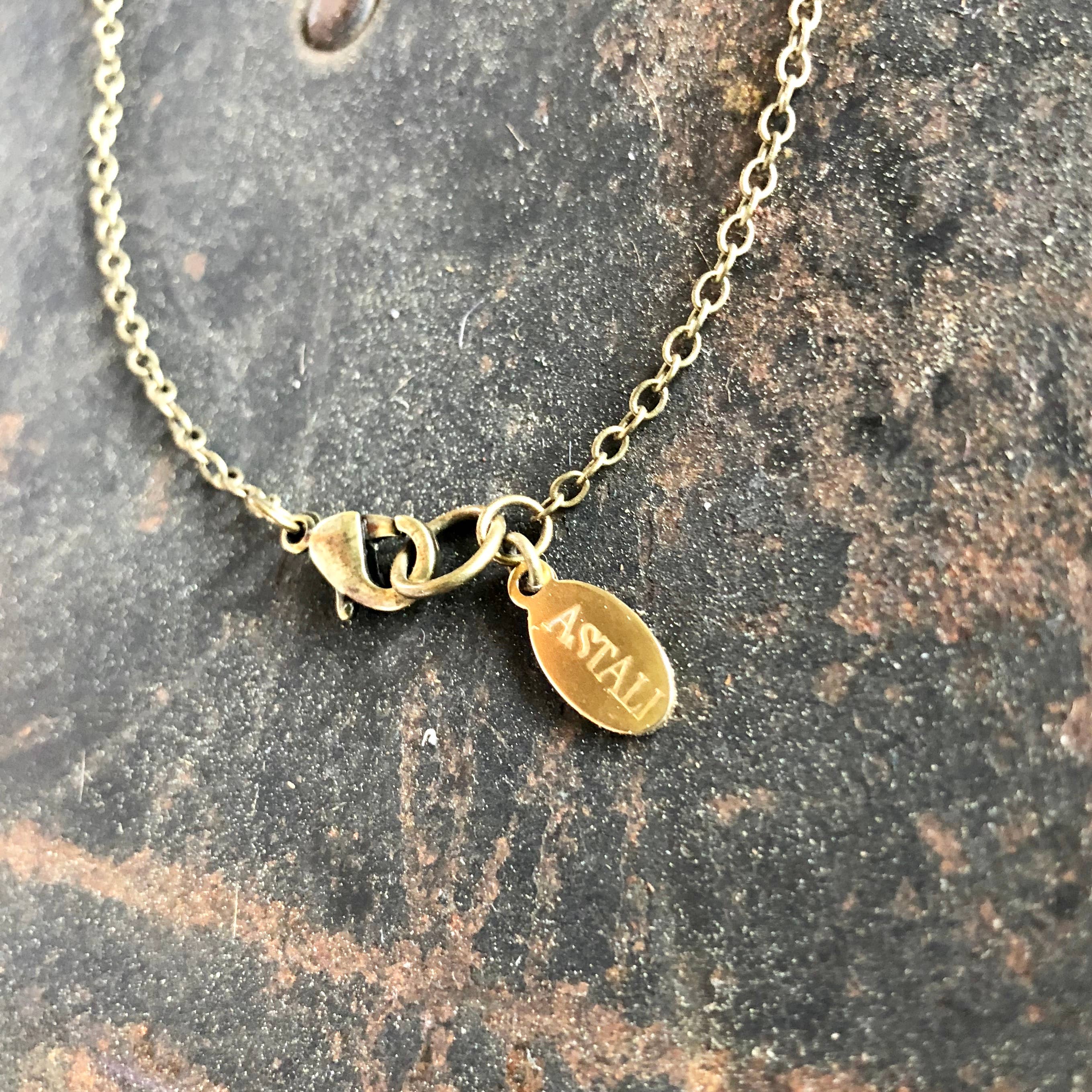 A close-up of the ASTALI Vintage Heads & Tails Peepshow Token Necklace’s gold clasp and engraved oval tag stands out against a textured, dark background, showcasing this collectible coin necklace’s unique appeal.