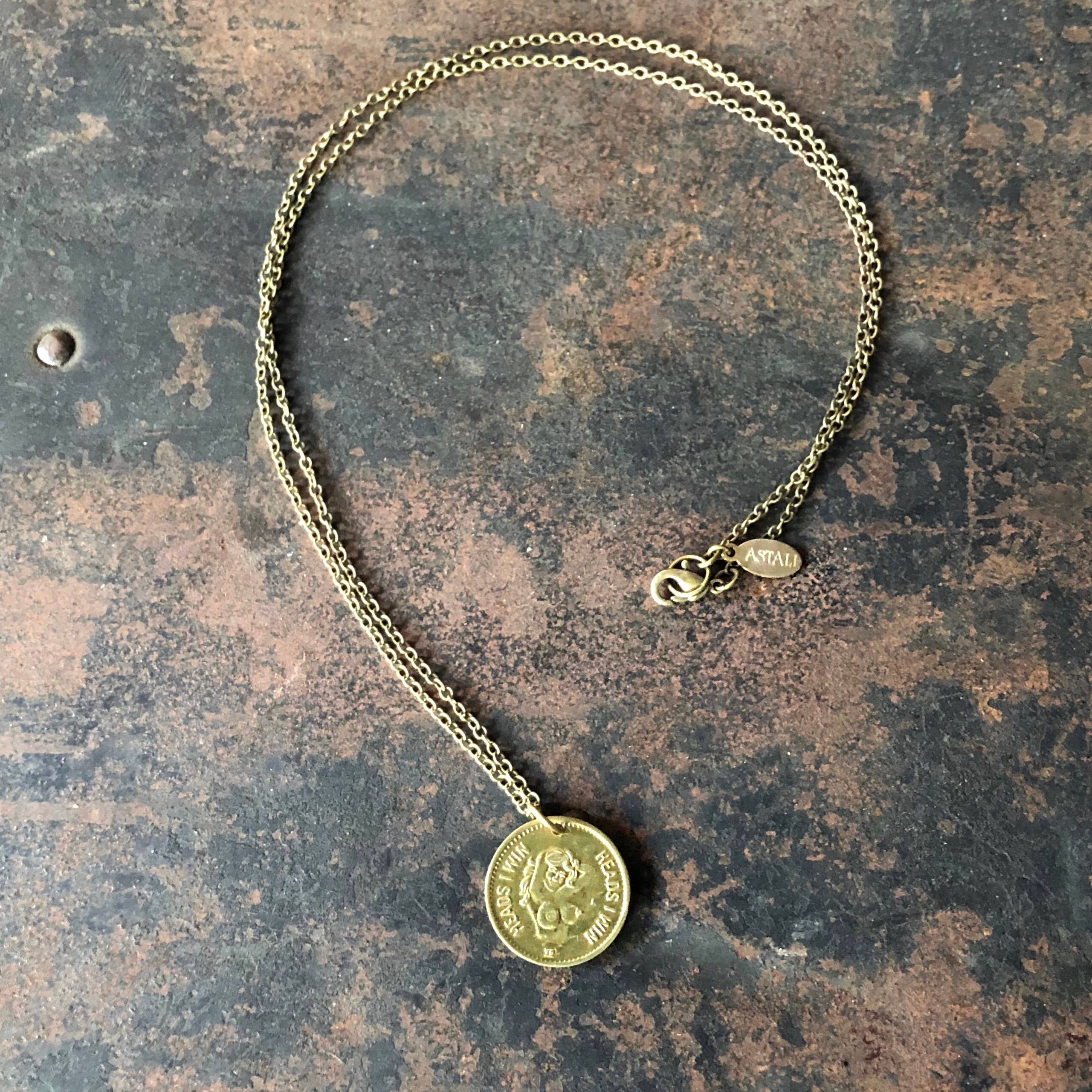 The ASTALI Vintage Heads & Tails Peepshow Token Necklace features a coin-like pendant with an engraved design and a small ITALY tag near the clasp, displayed on a weathered dark brown surface.