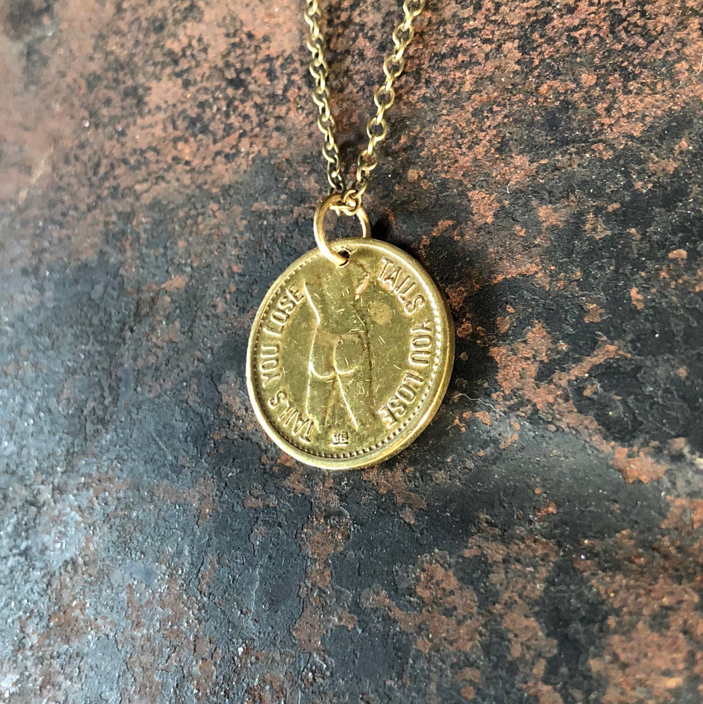 The ASTALI Vintage Heads & Tails Peepshow Token Necklace features a round, textured coin reading YOU LOSE, TAILS YOU LOSE with a backside image, displayed on a brown speckled stone surface.