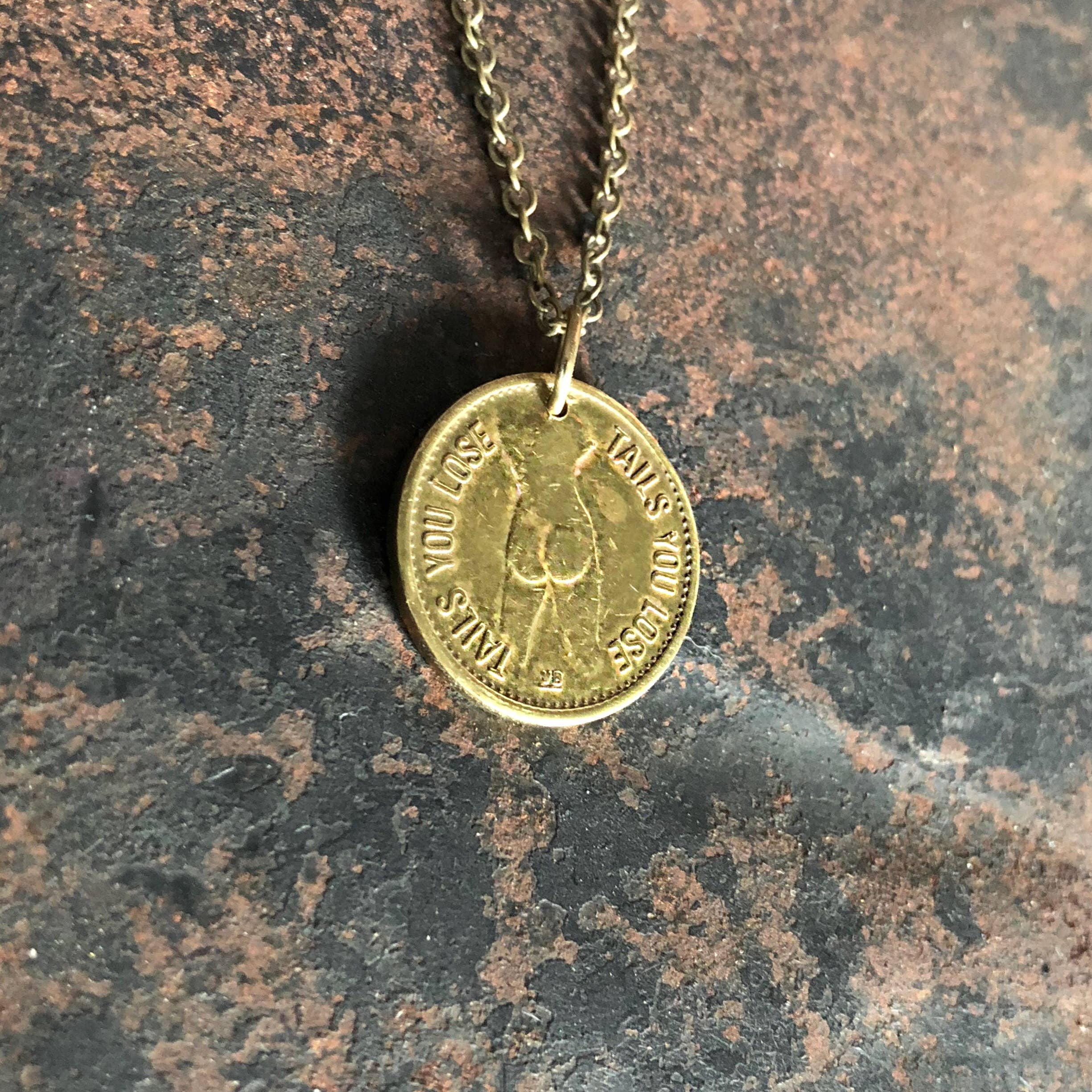 The ASTALI Vintage Heads & Tails Peepshow Token Necklace features a brass coin pendant inscribed HEADS YOU LOSE, TAILS YOU LOSE on an antique chain, shown resting on a textured surface—a unique collectible accessory.