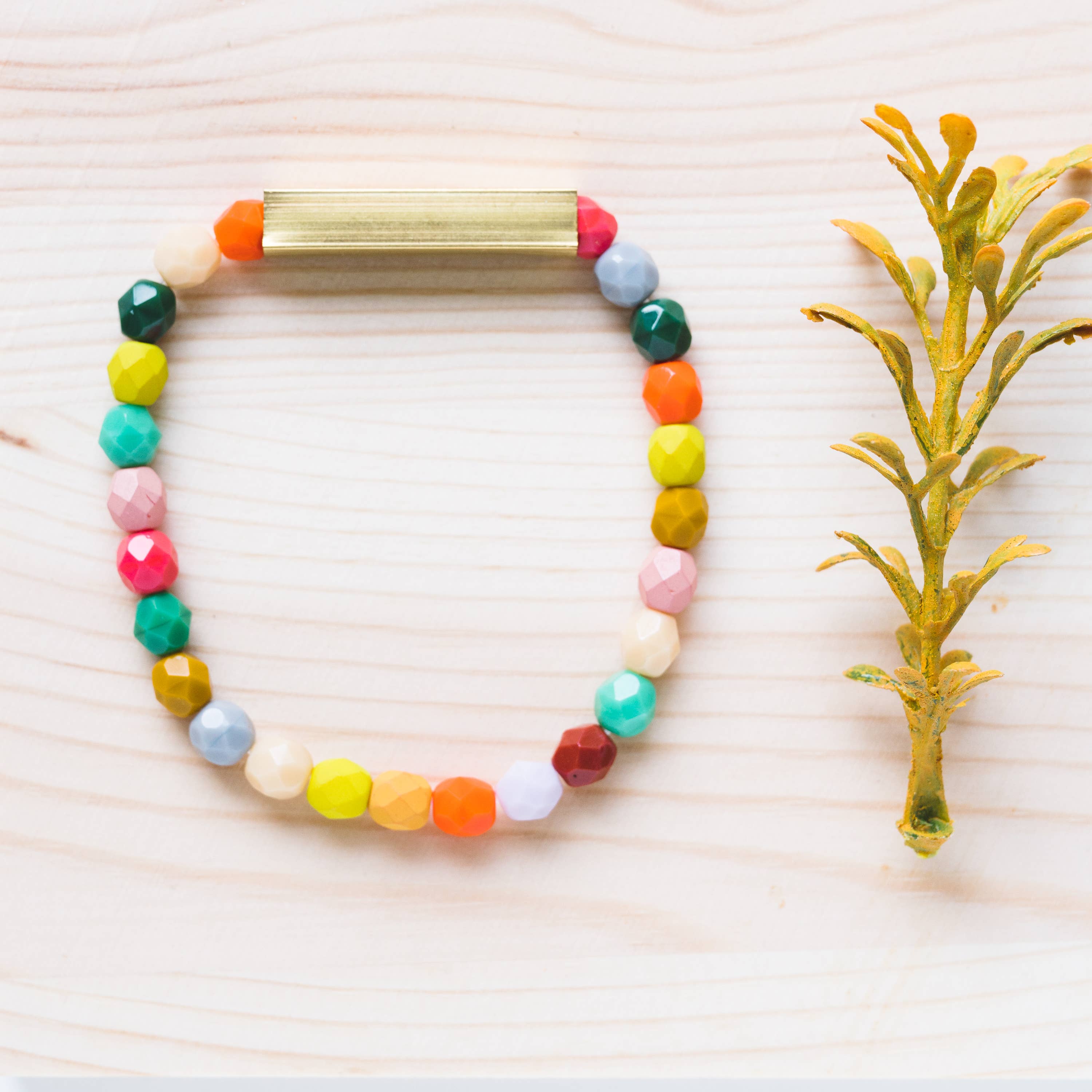 Mixed Beads and Brass Bracelet with colorful faceted beads and a sleek brass bar on light wooden background next to yellow sprig