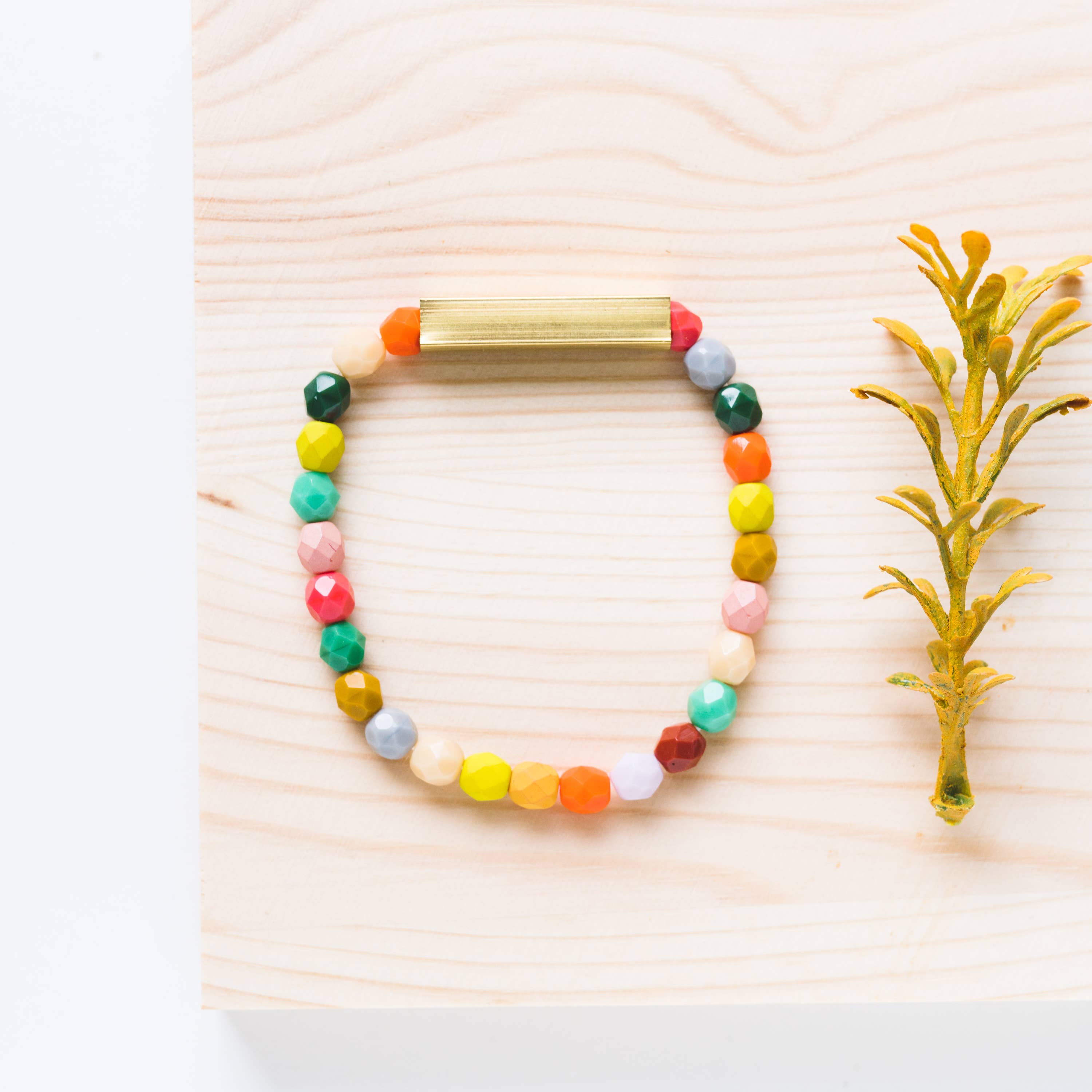 Mixed Beads and Brass Bracelet with colorful faceted beads laid on light wood next to a small yellow plant stem