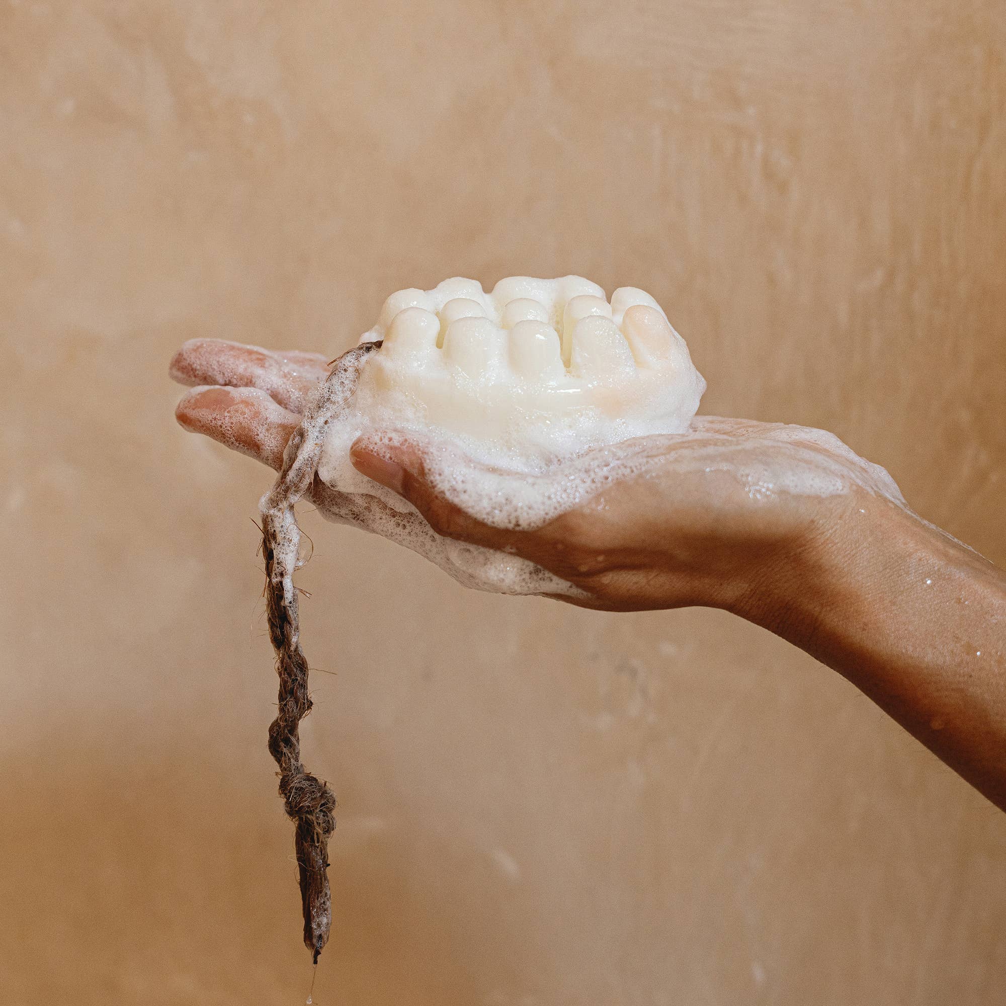 A hand holds a lathered Cedar and Myrrh Palo Santo + Shea Butter Bar Soap, shaped with rounded bumps and suds, a brown string hanging below—perfect for sensitive skin—set against a beige background.