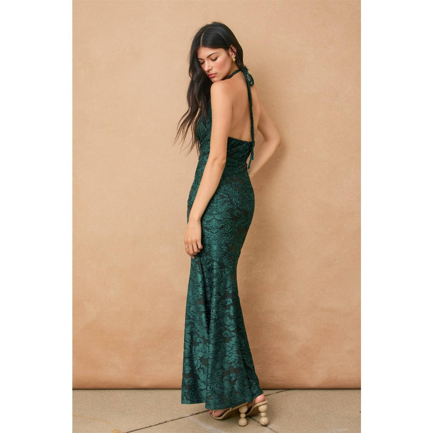 A woman poses sideways against a beige backdrop, wearing the Fame Draped Halter Neck Maxi Dress by Dress Forum—a fitted, floor-length evening gown in dark green with an open back and lace details, her long hair draped over one shoulder.