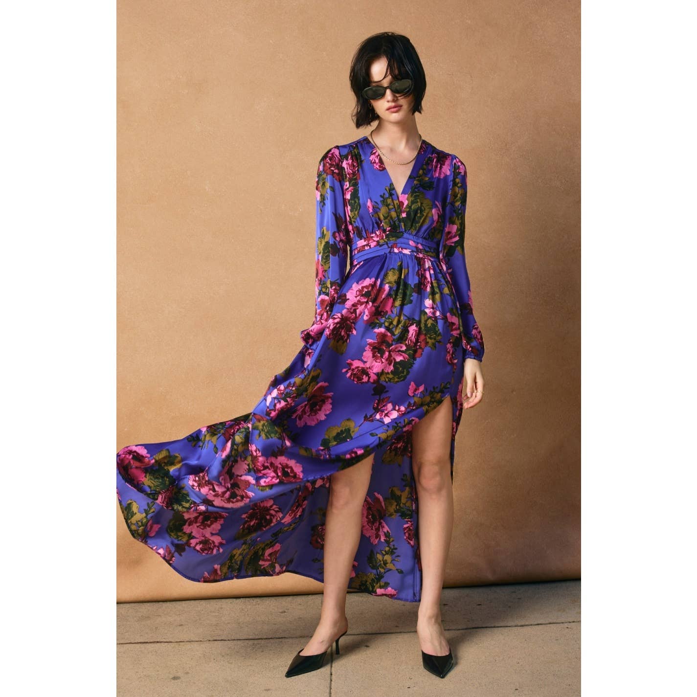 A woman in black sunglasses wears the Mesmerizing Essence Pleated Waist Maxi Dress by Dress Forum, featuring bold pink florals, flowing satin, long sleeves, pleated waist, side slit, and high-low hem. She pairs it with black pointed heels against a tan backdrop.