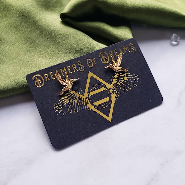 The Dreamers of Dreams Hummingbird 14k gold plated Stud Earrings are displayed on a black card with gold designs and “Dreamers of Dreams” in gold, set against green fabric and a white marble surface.