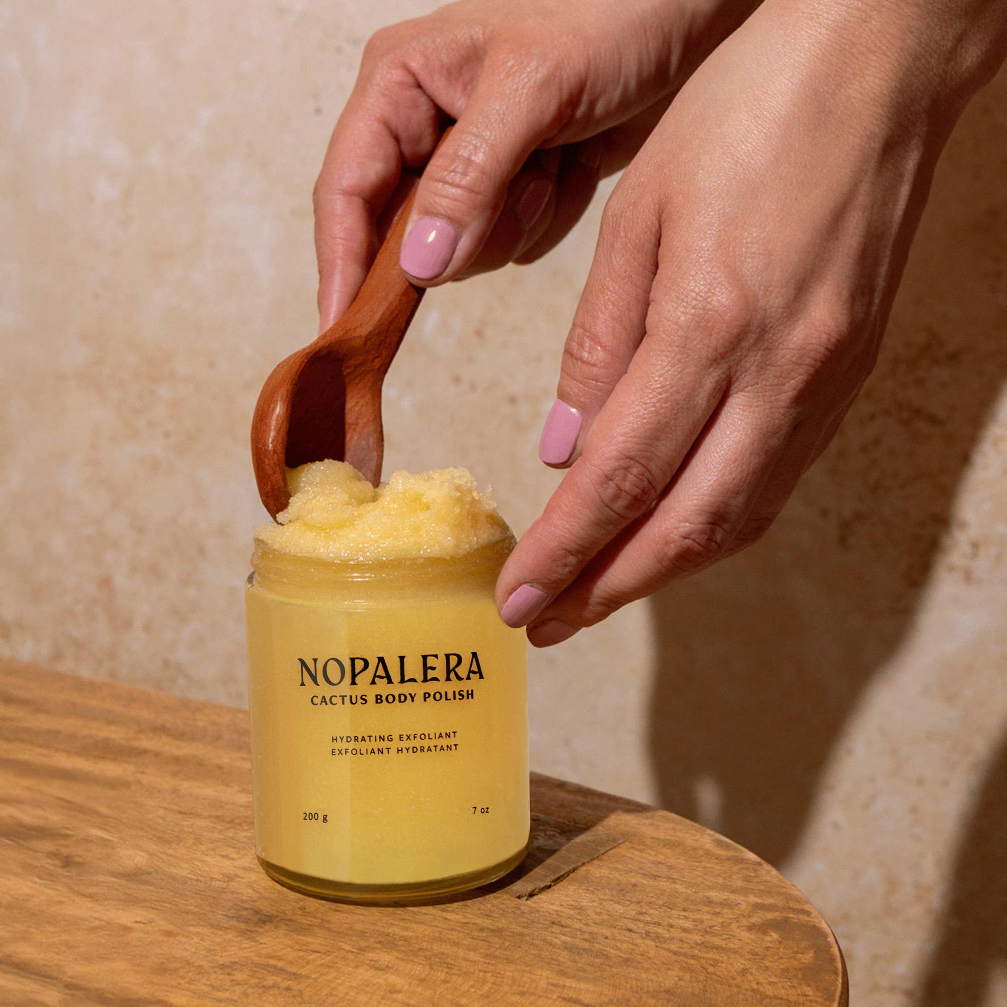 Hand scooping exfoliant from jar labeled Tangerine Cactus Body Polish with wooden spoon on wooden surface.