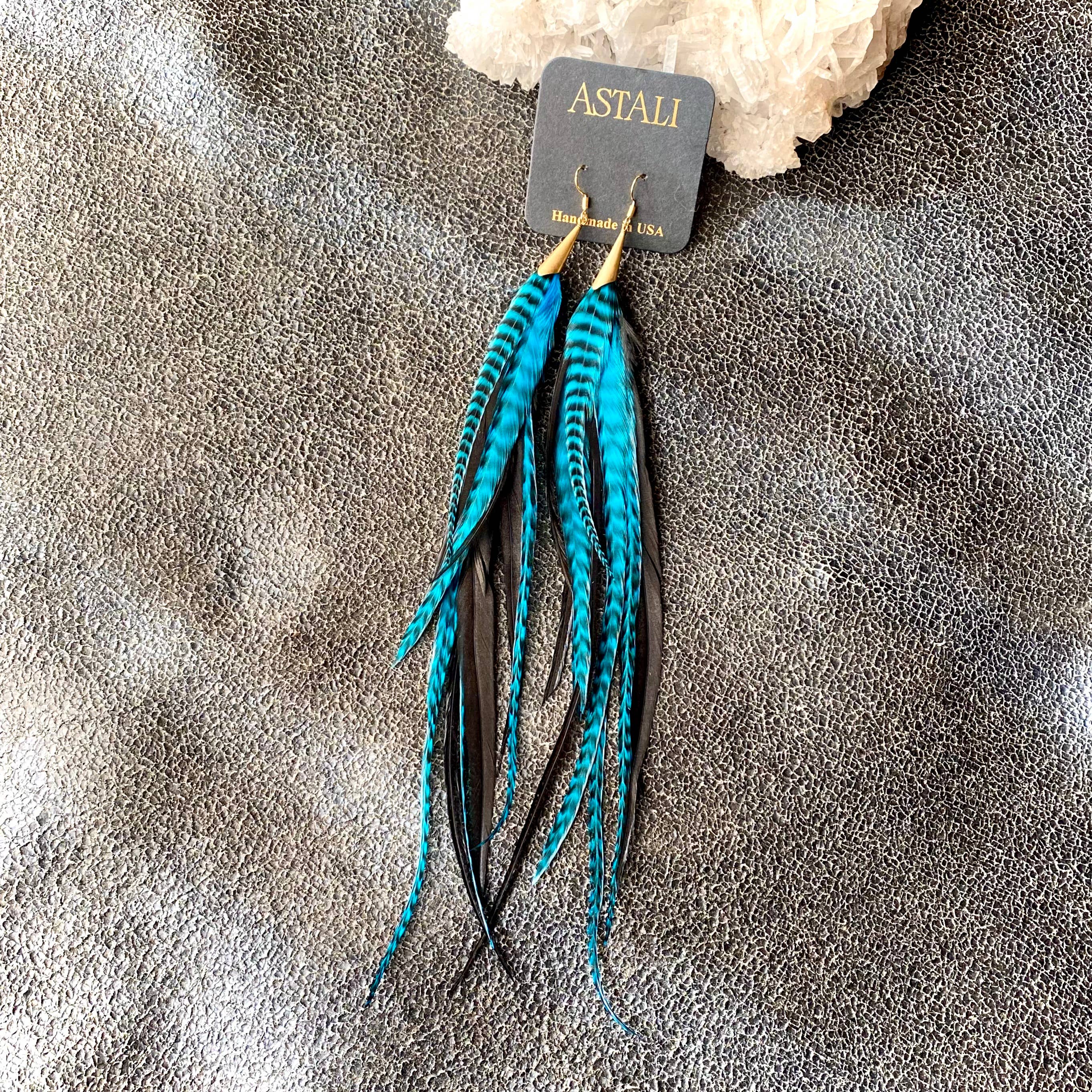 A pair of ASTALI Long Feather & Cone Earrings in Turquoise Grizzly & Black/Gold, featuring sustainably sourced feathers and gold hooks, displayed on a branded ASTALI Handmade in USA card against a textured gray background.