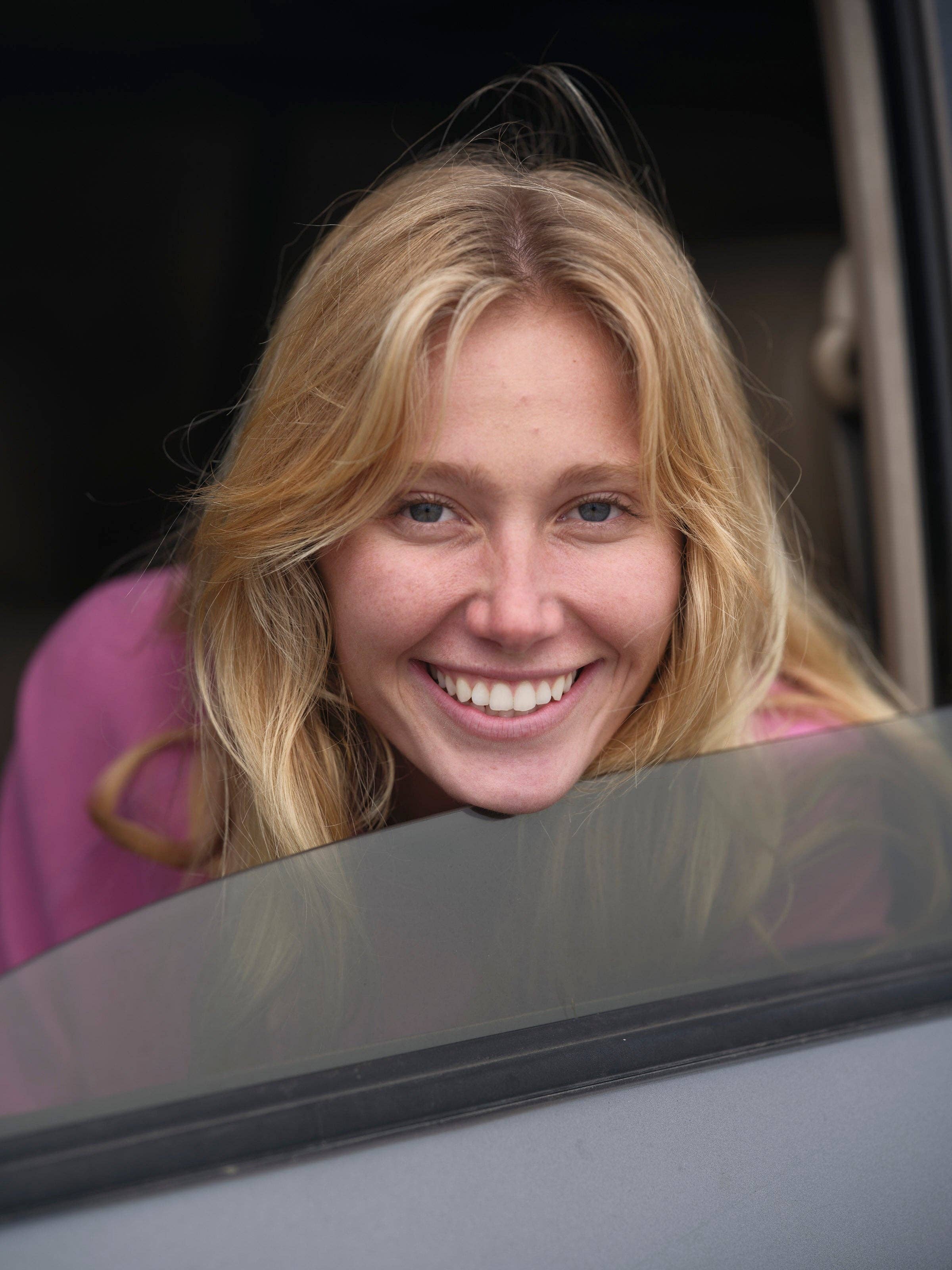 A young woman with long blonde hair smiles brightly while leaning out of a car window, wearing the pink Mollusk Visions Crew sweatshirt.