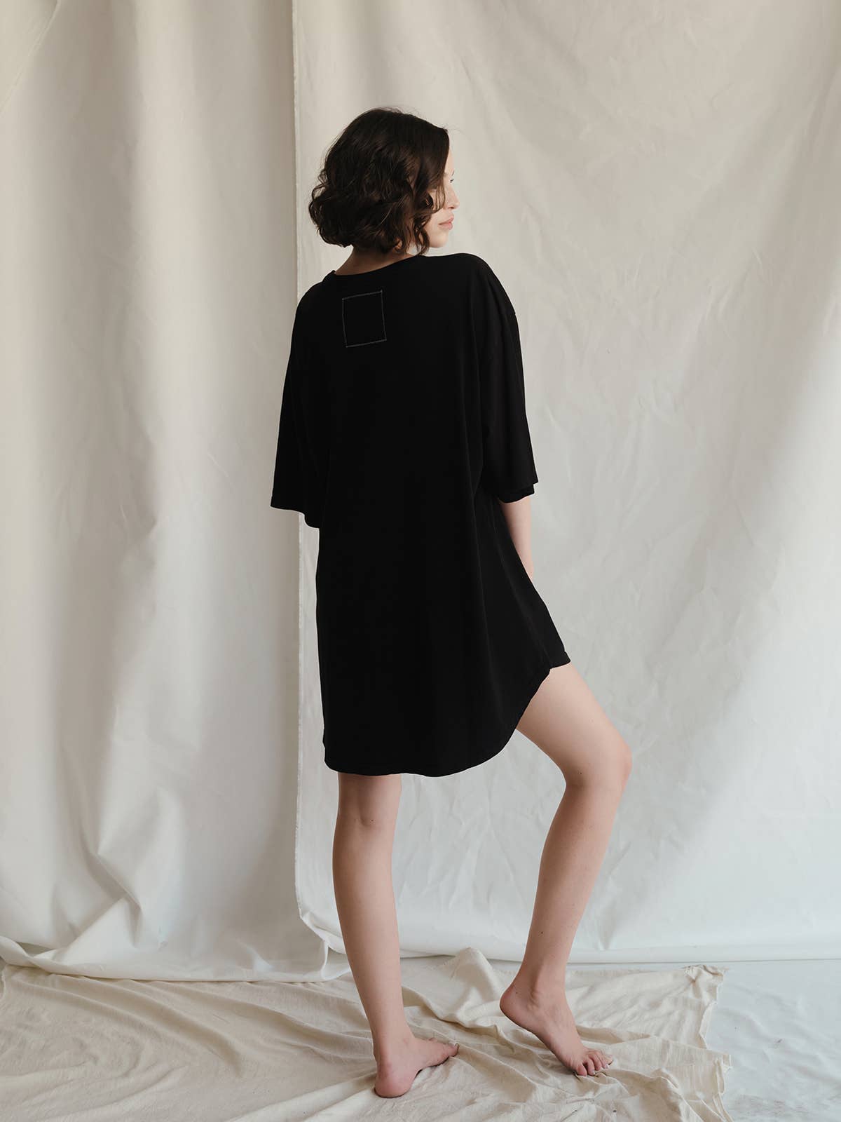 A woman with short curly hair stands barefoot on draped fabric, modeling the Blondie Camp Oversized Tee by People Of Leisure. Ethically made from recycled cotton, the loose black tee is shown from the back against a light, textured backdrop.