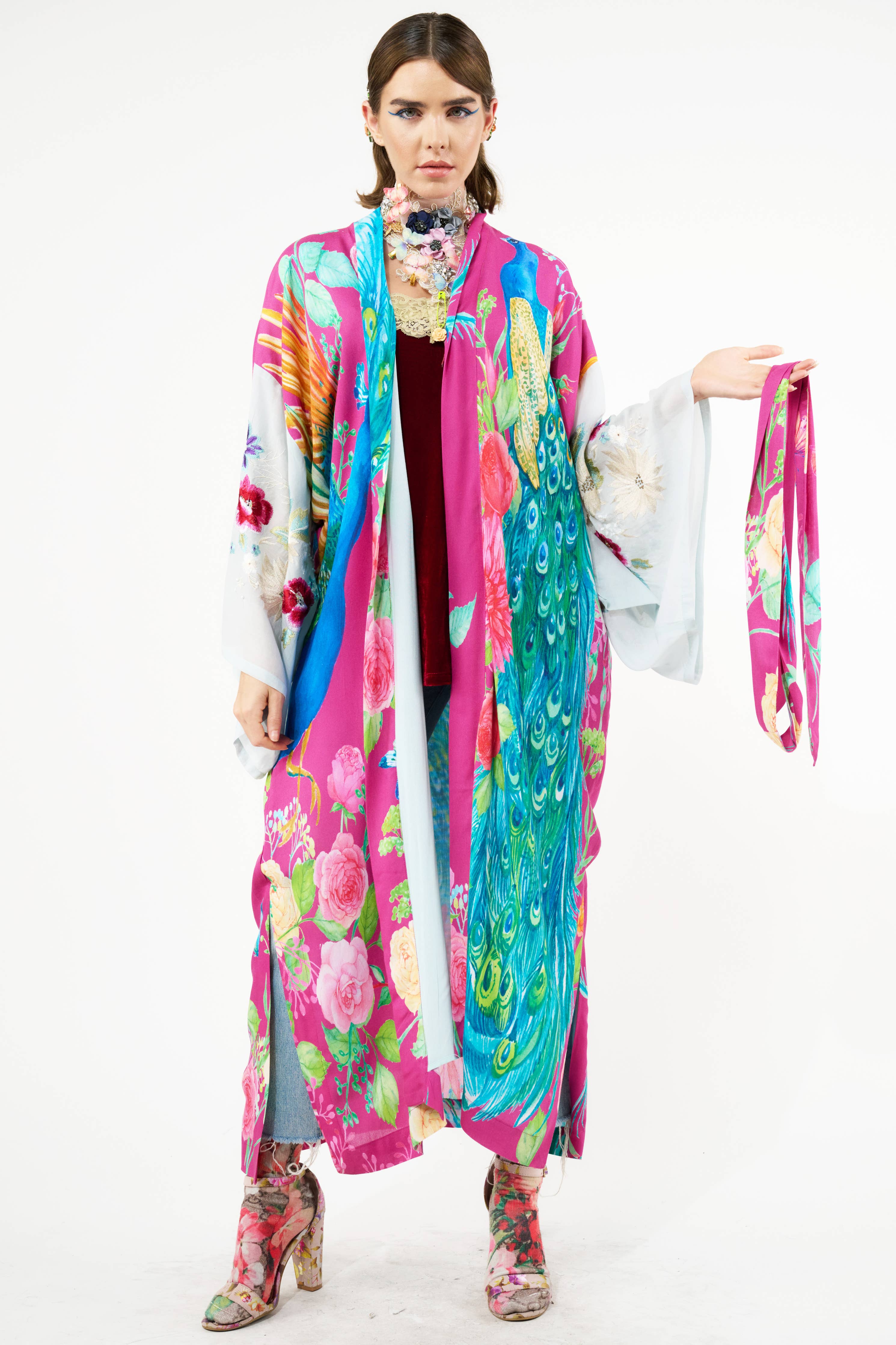 A woman stands against a white background wearing Aratta’s Marie Antoinette Kimono with wide, embroidered floral sleeves, blue jeans, and matching boots. She holds a coordinating sash—a perfect bohemian statement layer.