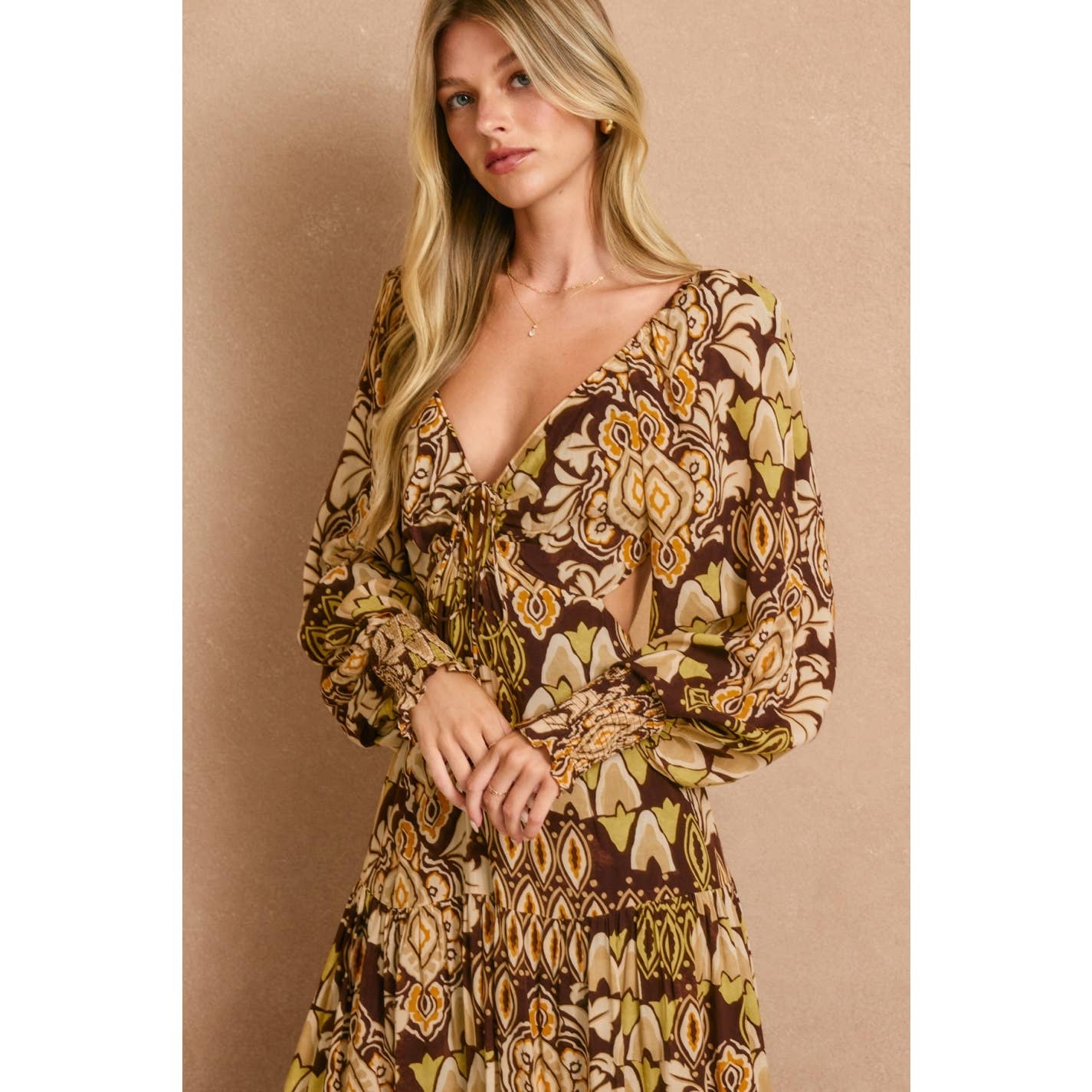 A woman with long blonde hair wears the Dress Forum Stone Ember Keyhole Long Sleeve Maxi Dress in brown, yellow, and beige tones with a deep V-neck, posing against a beige background.