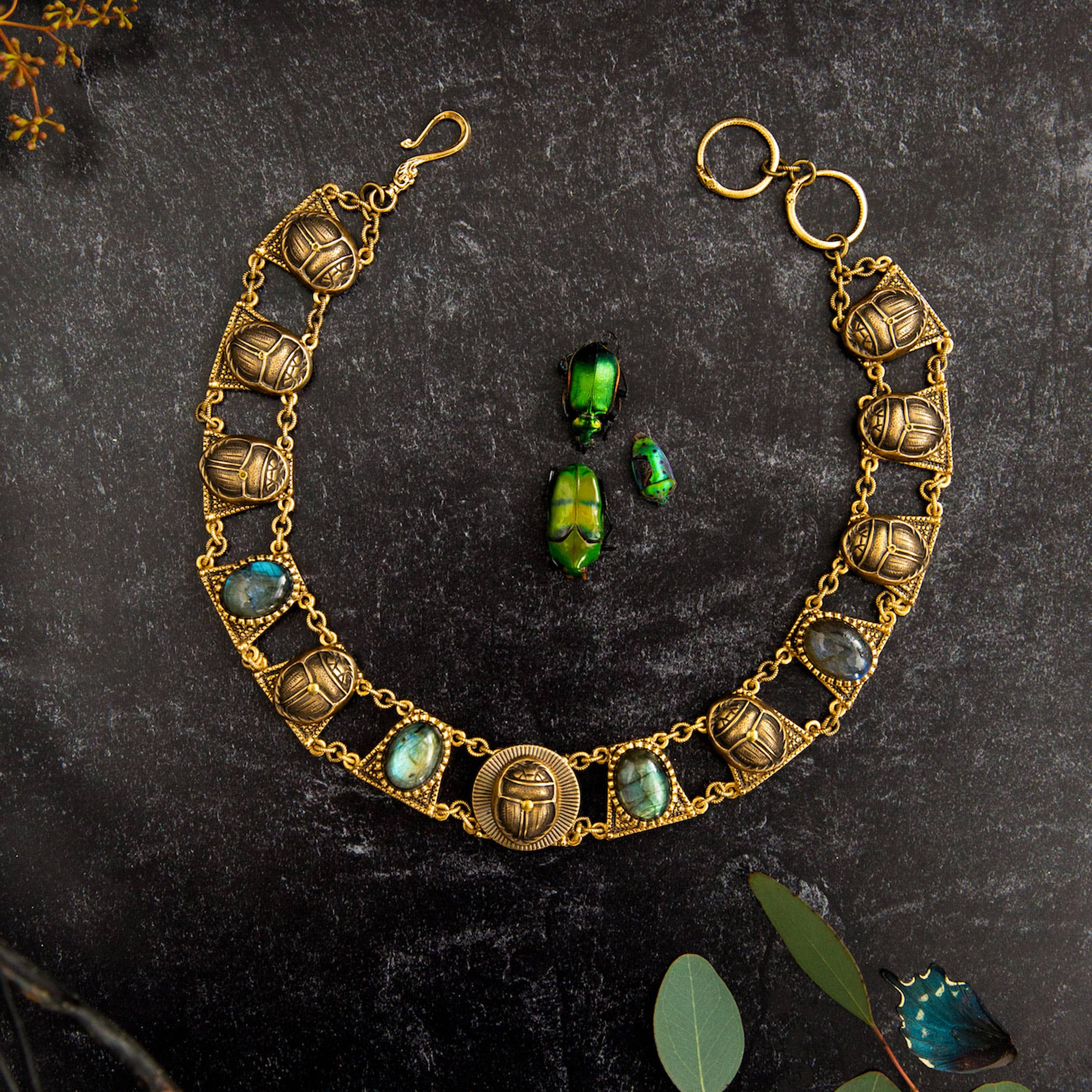 Sacred Scarab Collar Necklace with brass scarab charms and iridescent stones on a dark textured background