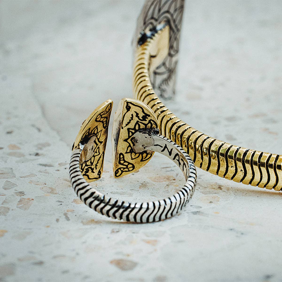A close-up of two intricately designed bahgsu jewels rings: one is the gold and black adjustable Natural Mystic Ring - Moonstone with engraved patterns, the other a sterling silver ring with black accents and a unique split open design on a speckled surface.