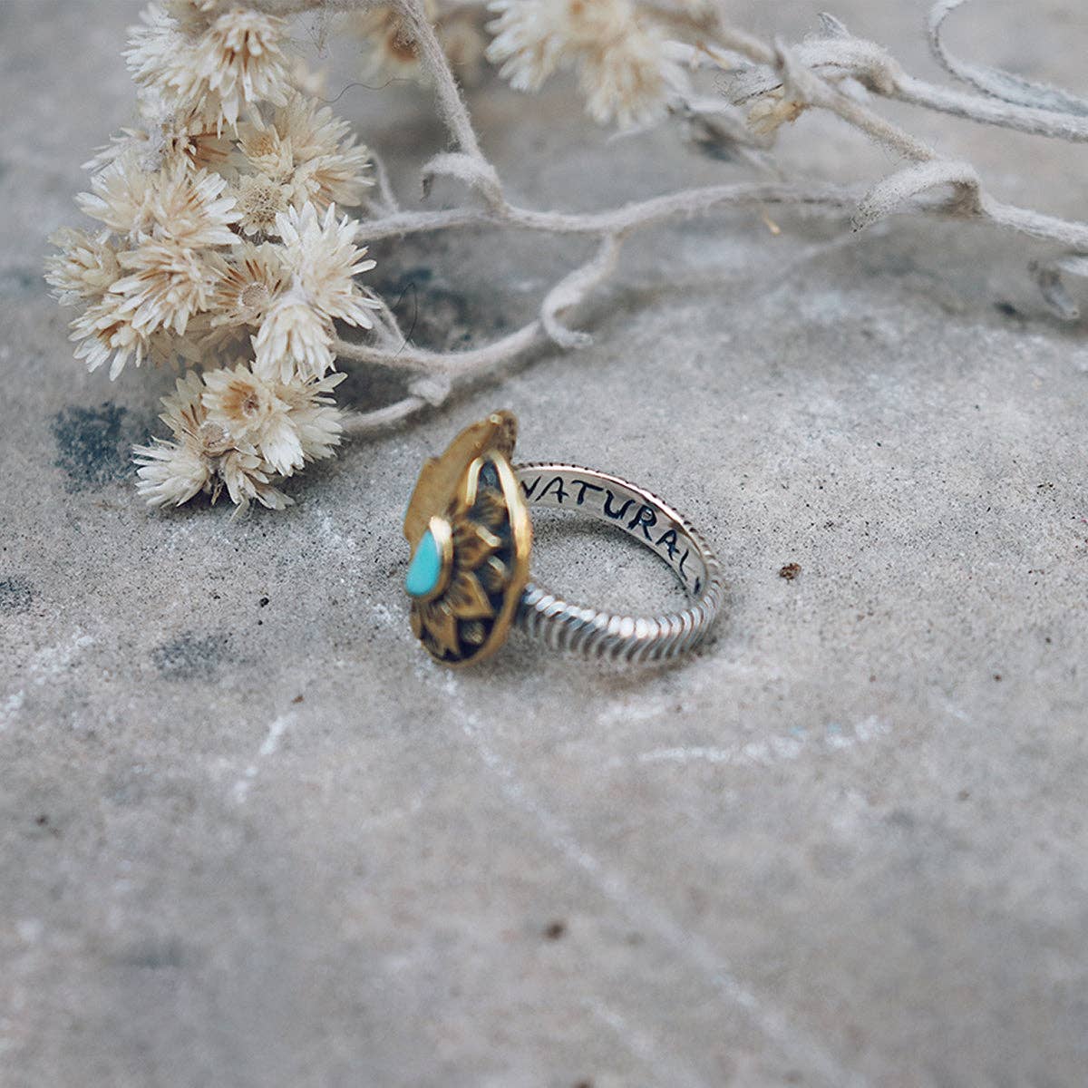 The bahgsu jewels Natural Mystic Ring - Turquoise features a sterling silver band with a gold sun-shaped design and turquoise center, engraved with NATURAL inside, displayed on concrete next to small dried white flowers.