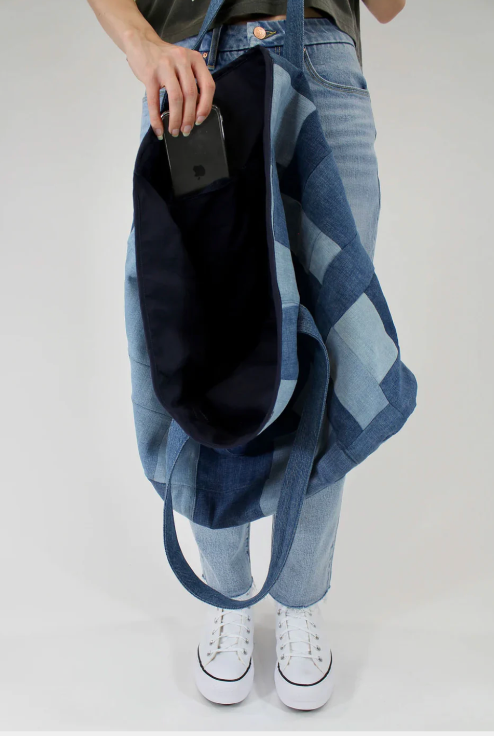 A person in light blue jeans and white sneakers holds the Porter Blue Apparel PATCHWORK TOTE, placing a black smartphone into its spacious interior pocket. The background is plain and light-colored.