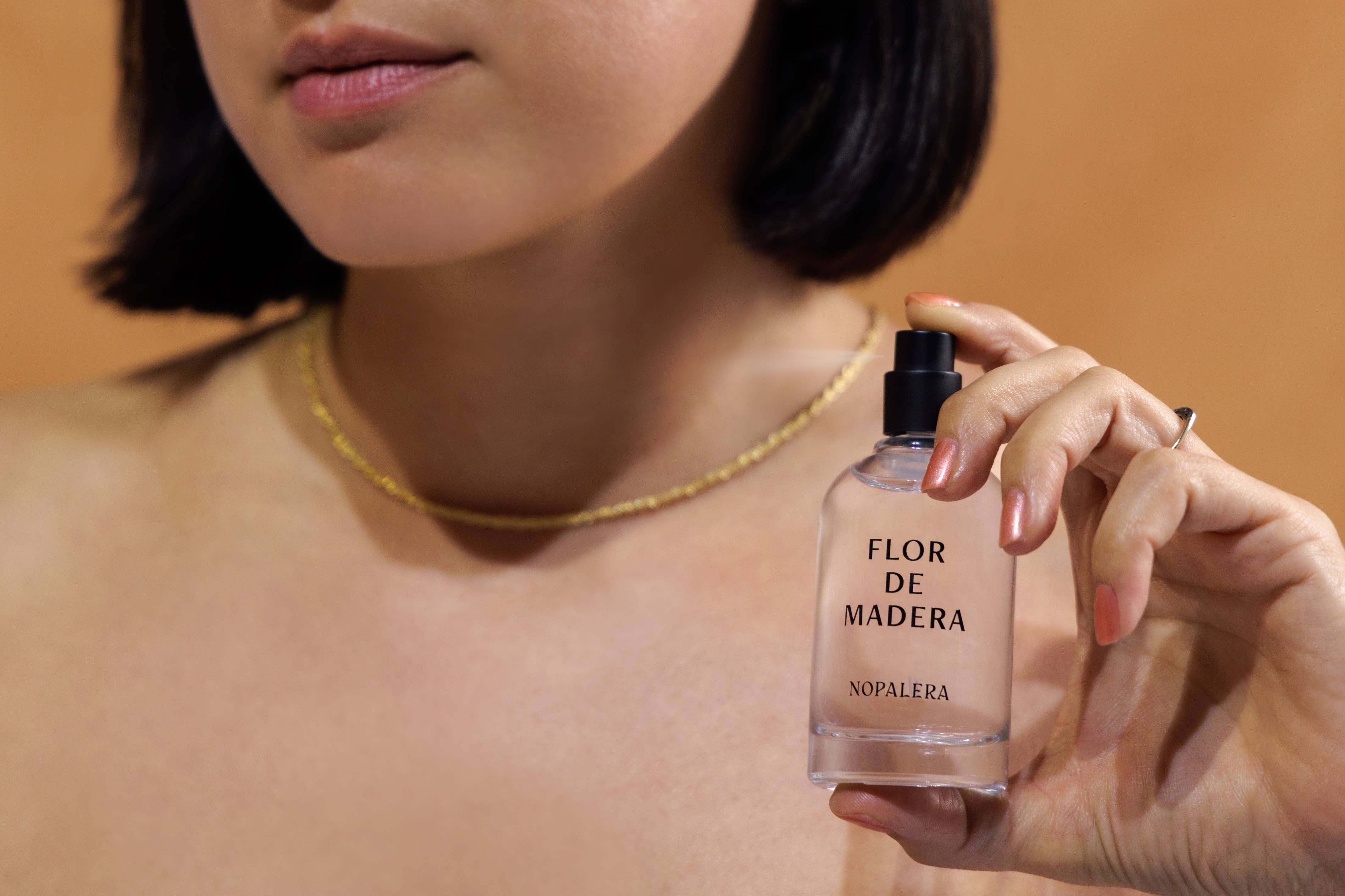 A person with short dark hair holds the Nopalera Flor de Madera Eau de Parfum – 10mL Travel Spray near their face, wearing a thin gold necklace. The neutral background highlights this clean scent inspired by Mexican citrus notes.