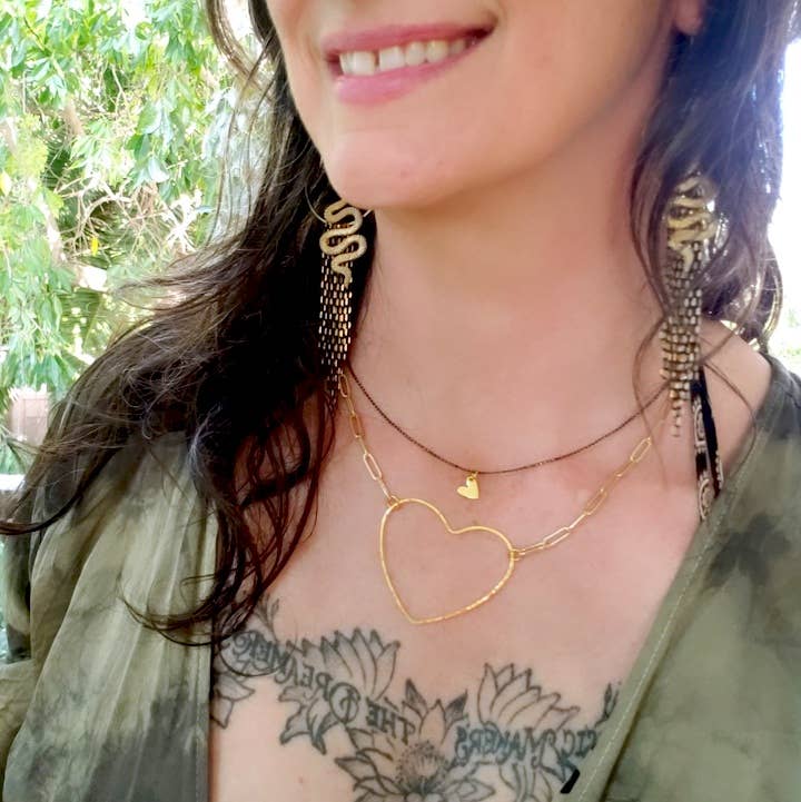 A woman smiles outdoors in a green top, wearing Dreamers of Dreams Big Gold Heart Paperclip Chain Necklace and large gold earrings. A chest tattoo is visible above the neckline, with blurred trees in the background.