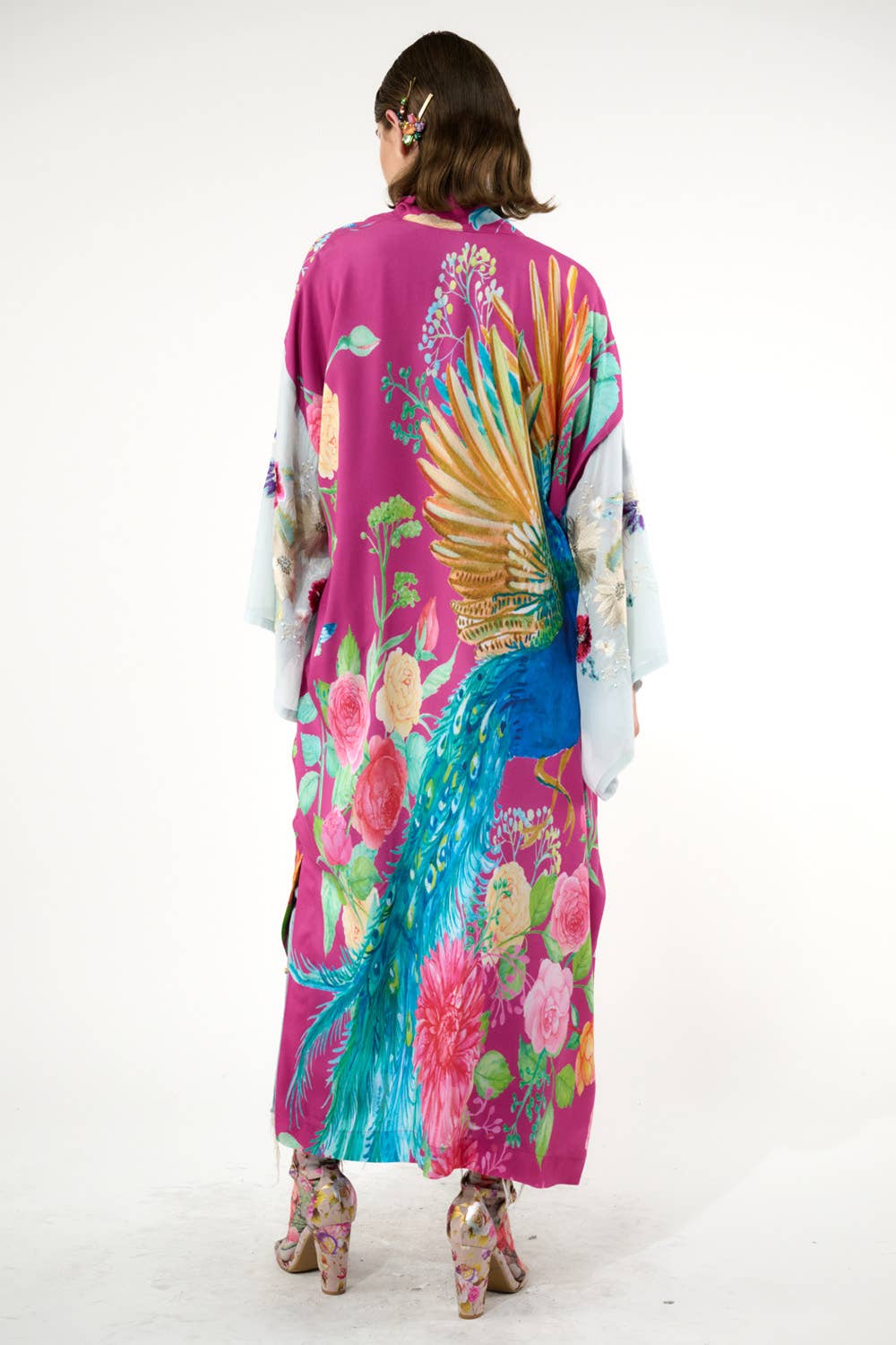 A person faces away, wearing the Marie Antoinette Kimono by Aratta with embroidered floral sleeves. Their wavy hair is clipped back, and they wear high-heeled shoes adorned with a floral pattern—a striking statement look.