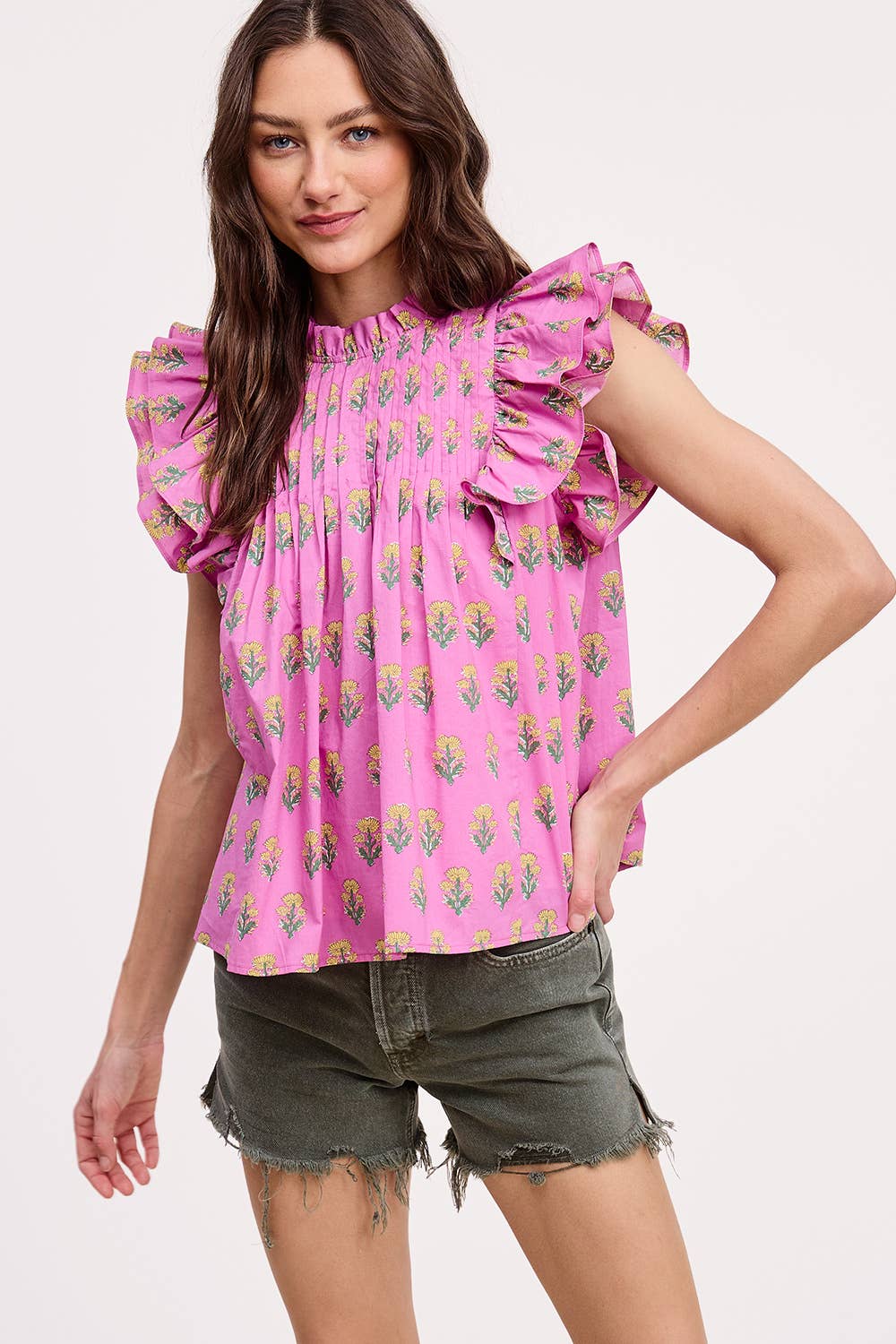 A woman smiles against a plain light background, wearing Ultramarines Celeste Flutter Sleeve Blouse in pink with a floral pattern and green frayed denim shorts, one arm bent with her hand on her hip.