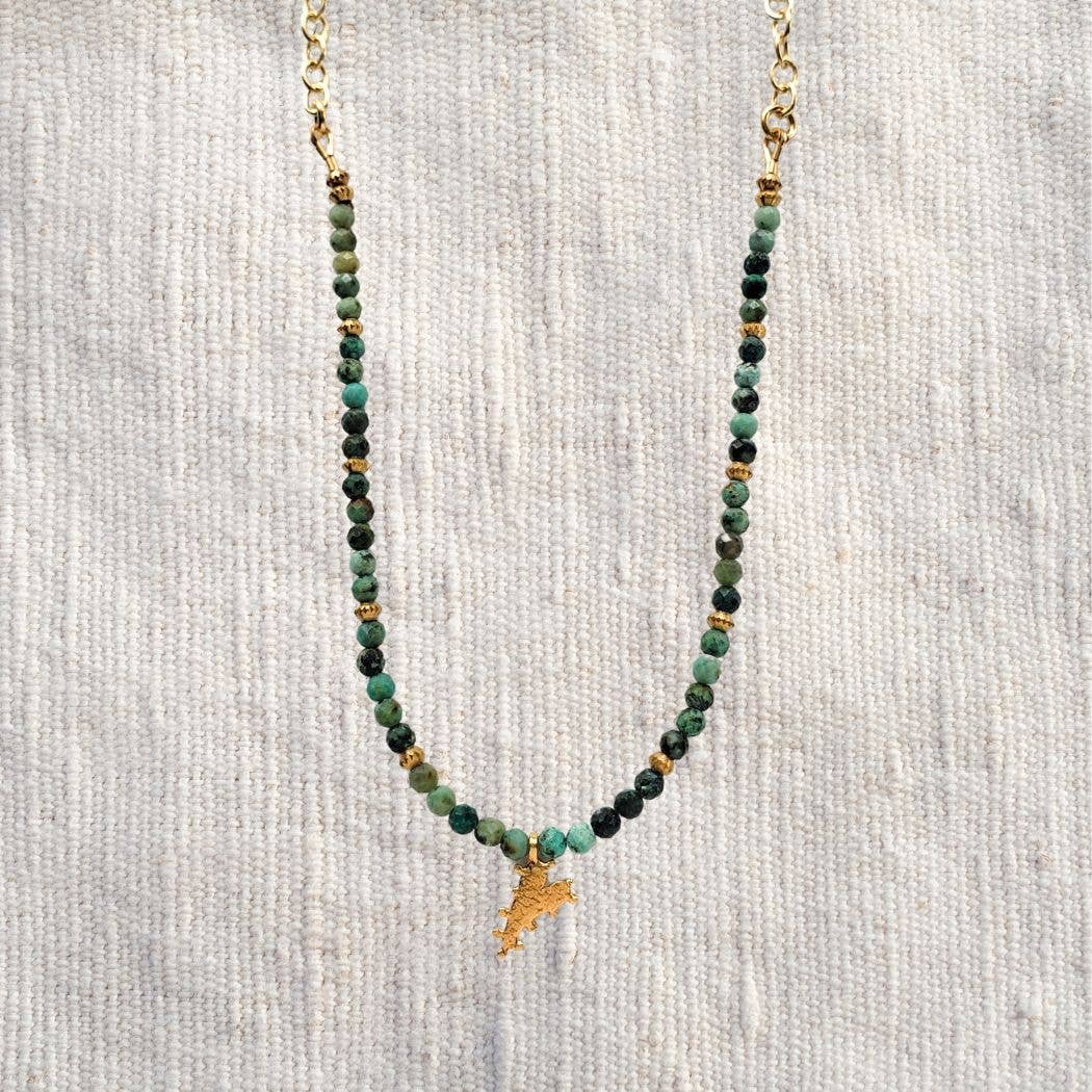 The Chrysocolla Beaded Heart Charm Necklace by Dreamers of Dreams features green and gold beads and a heart charm, displayed on cream fabric—a bold bohemian accessory.