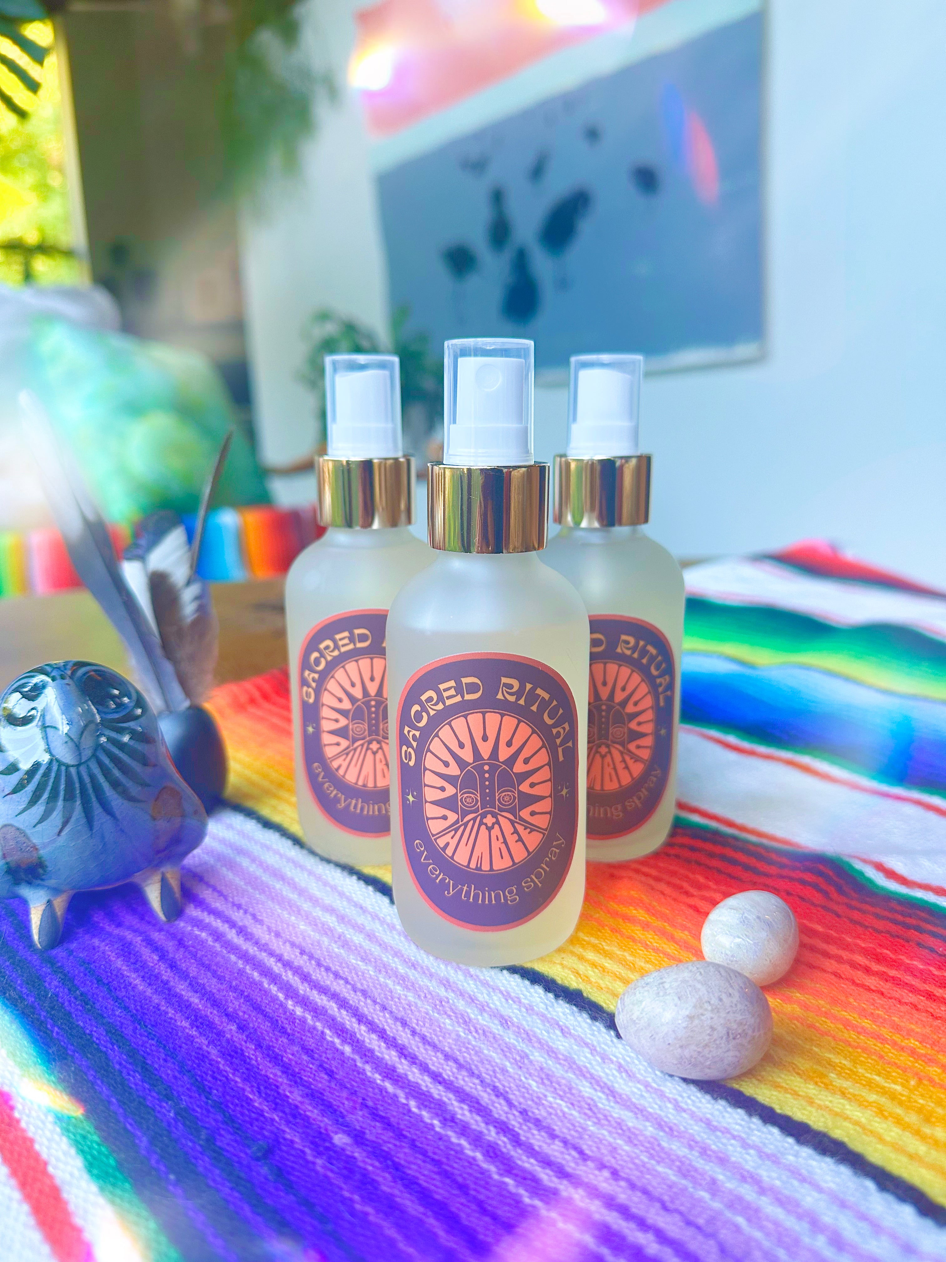 Three frosted glass bottles of HUM+BEE’s Sacred Ritual Everything Spray—a versatile mist for home and body—sit on a vibrant rainbow-striped cloth, accompanied by a ceramic bird, two stones, and a feather in the sunlight.