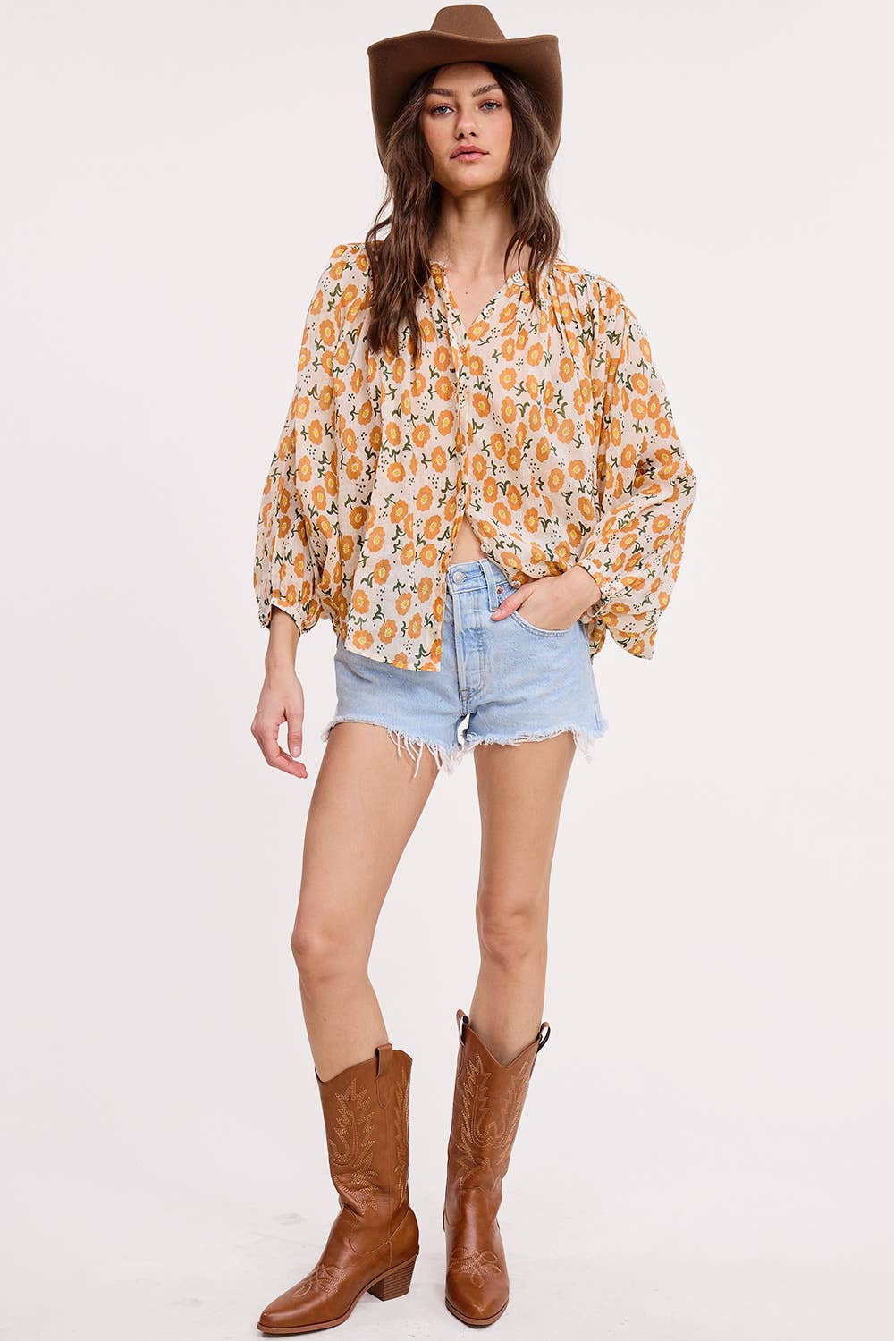 A woman wearing the Ultramarine Meadow Breeze Linen Blouse with yellow floral prints, balloon sleeves, light blue denim cutoff shorts, a brown cowboy hat, and brown cowboy boots stands against a plain white background.
