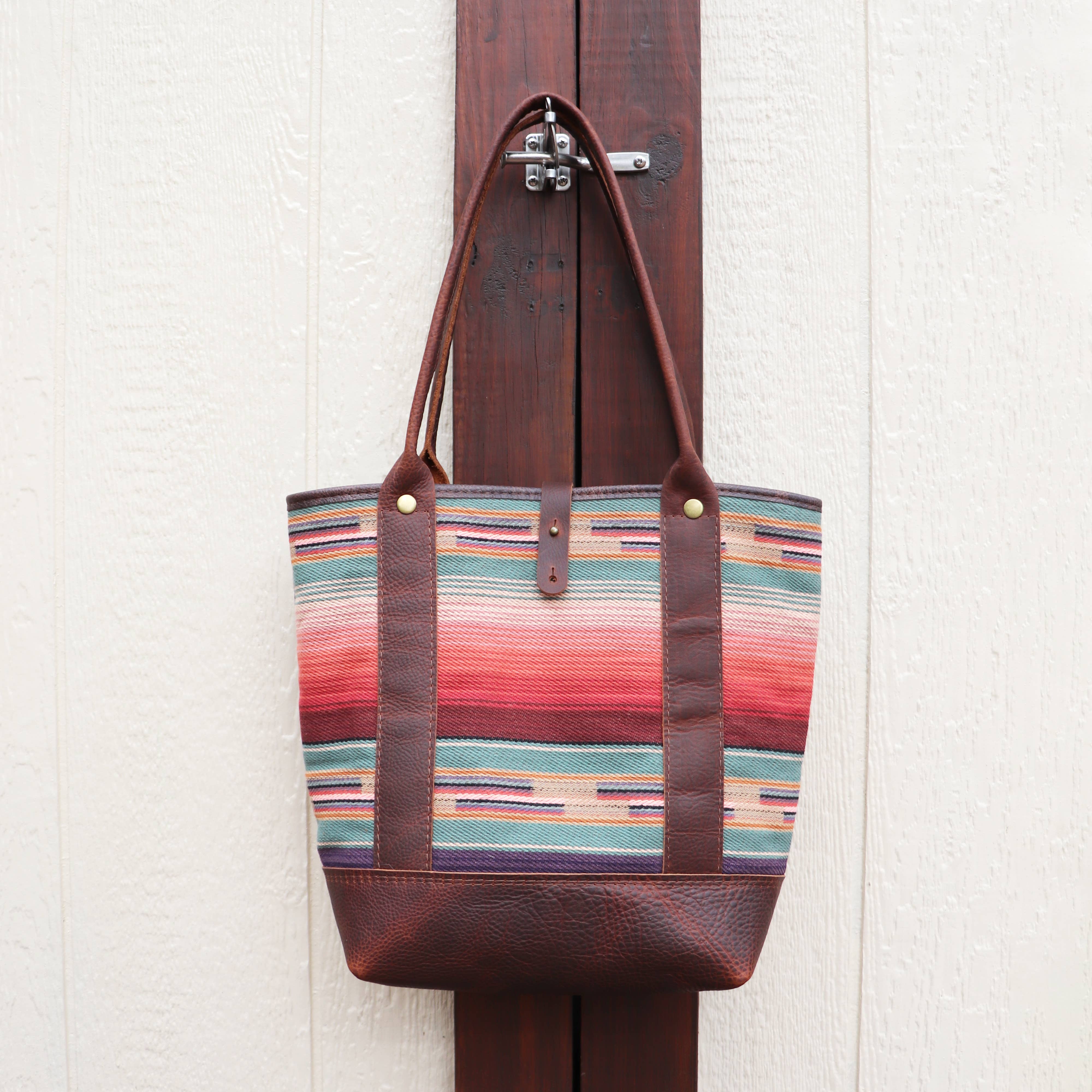 The Pearl Serape Tote by Hawks & Doves, a colorful handmade bag of striped serape fabric with brown leather accents, hangs on a wood panel against a light textured wall.
