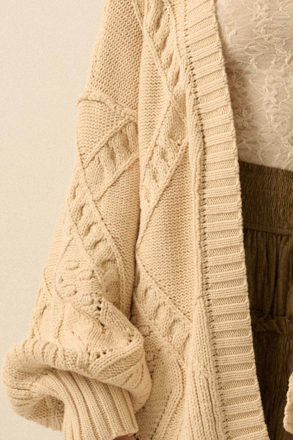 A close-up shows a person in a Promesa USA Cable-Knit Oversize Balloon-Sleeve Cardigan with beige textured knit and diamond pattern, layered over a white lace top and brown pleated skirt. The cardigan sleeve is slightly rolled up.