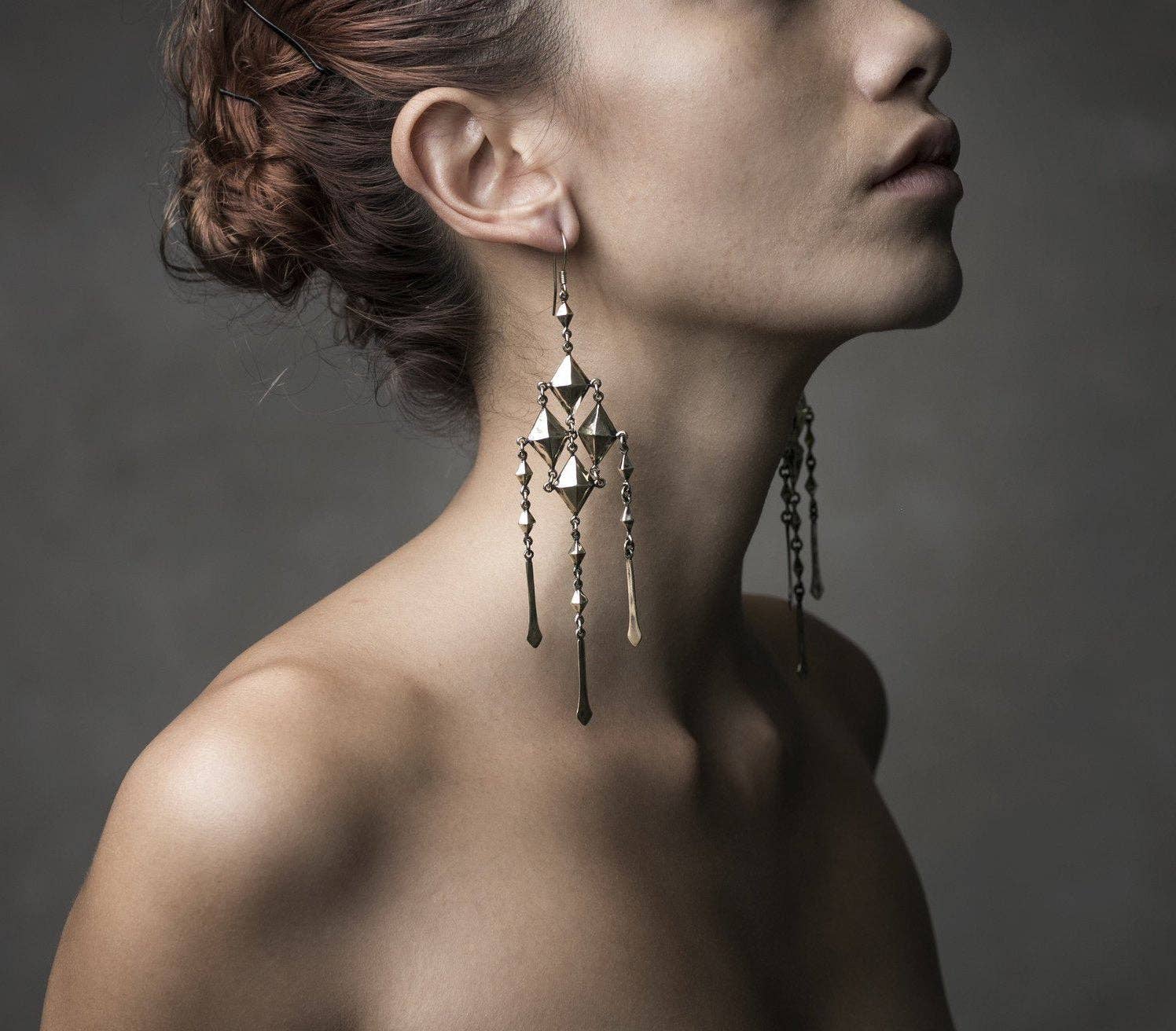 A woman with her hair in an updo models the Jan Hilmer + Sparrow Gem chainmail earrings—geometric brass dangles with hand cast chains and sterling silver hooks—her face partially in profile against a dark, neutral background.