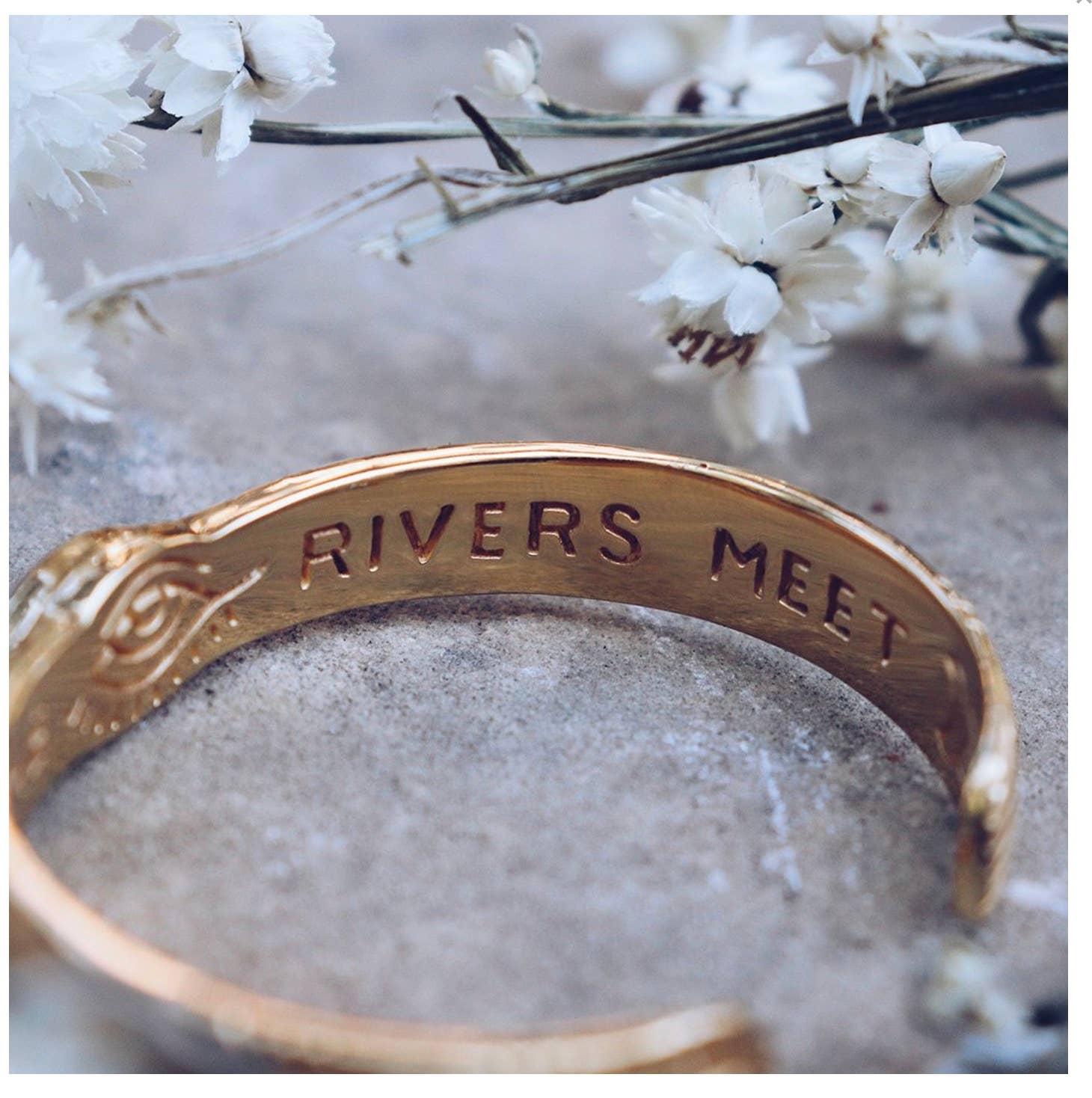Three Rivers Cuff in brass with engraved message surrounded by delicate white flowers on stone surface