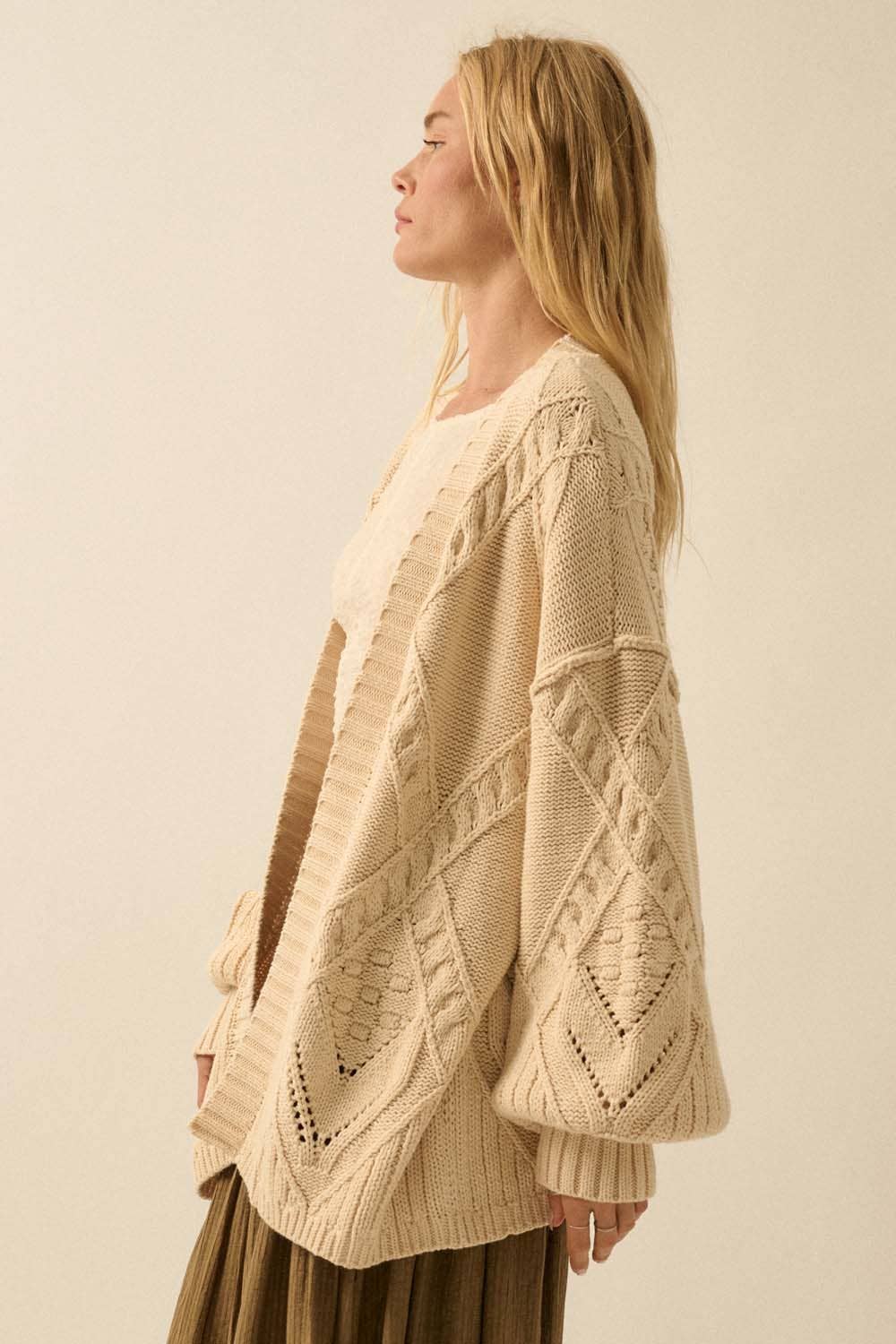 A woman with long blonde hair is shown in profile against a neutral background wearing the Promesa USA Cable-Knit Oversize Balloon-Sleeve Cardigan in a textured, cream-colored knit.