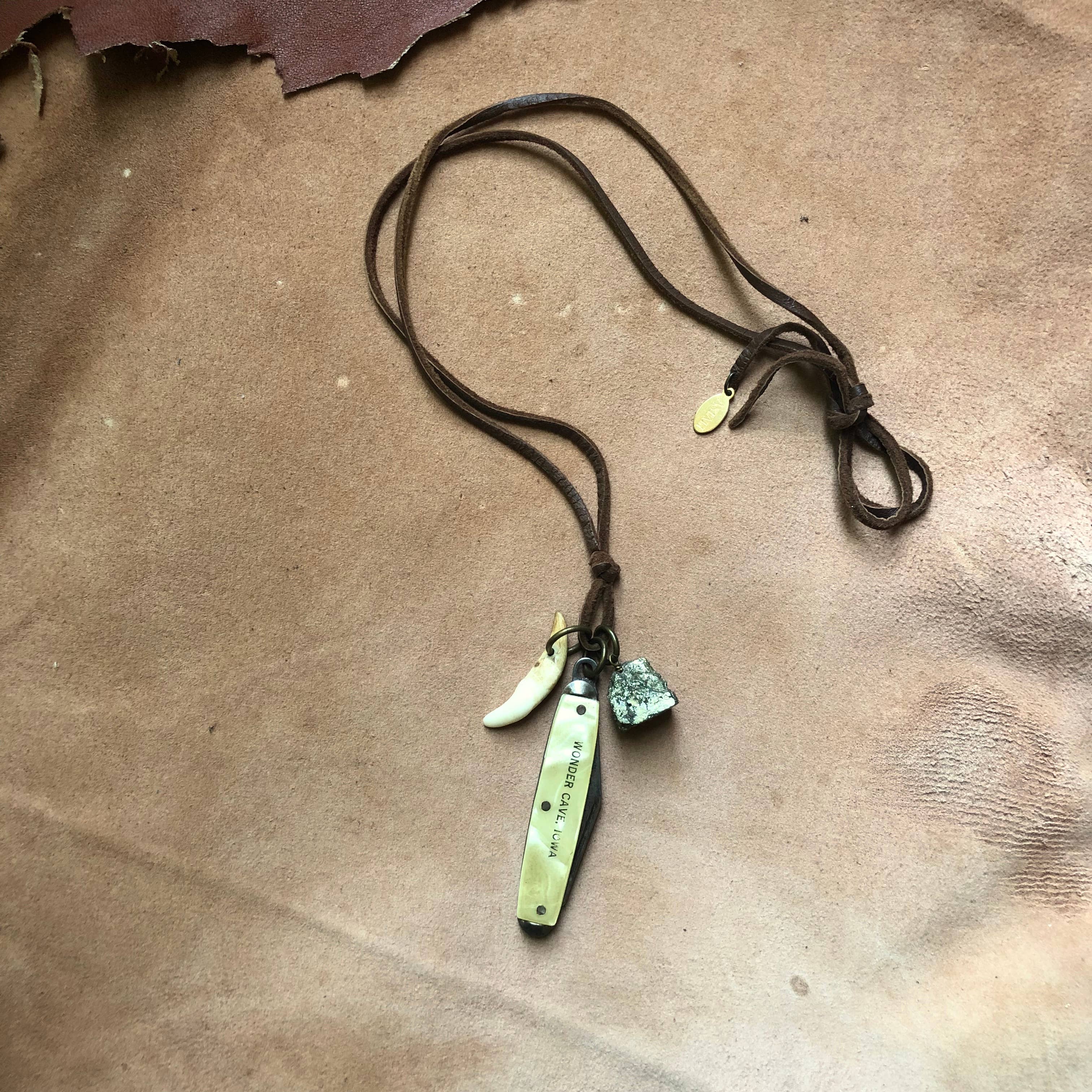 The ASTALI Vintage Pocket Knife & Coyote Tooth Necklace features deerskin leather, a brass tag, coyote tooth-inspired charm, and pyrite stone, displayed on tan leather.