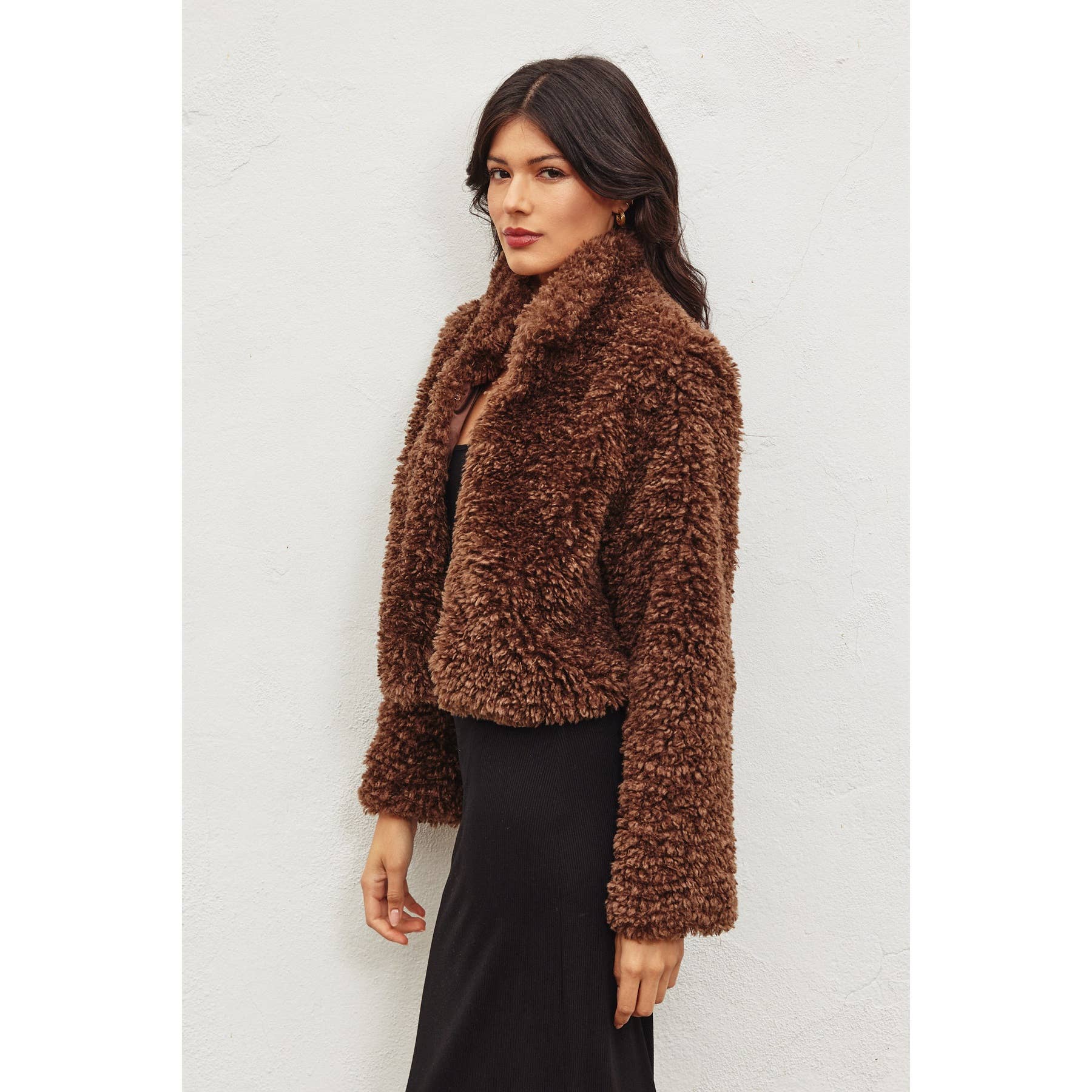 A woman with long dark hair wears the Dress Forum Soft Teddy Open Front Cropped Jacket in brown over a black dress, standing against a white wall and looking at the camera with a neutral expression.