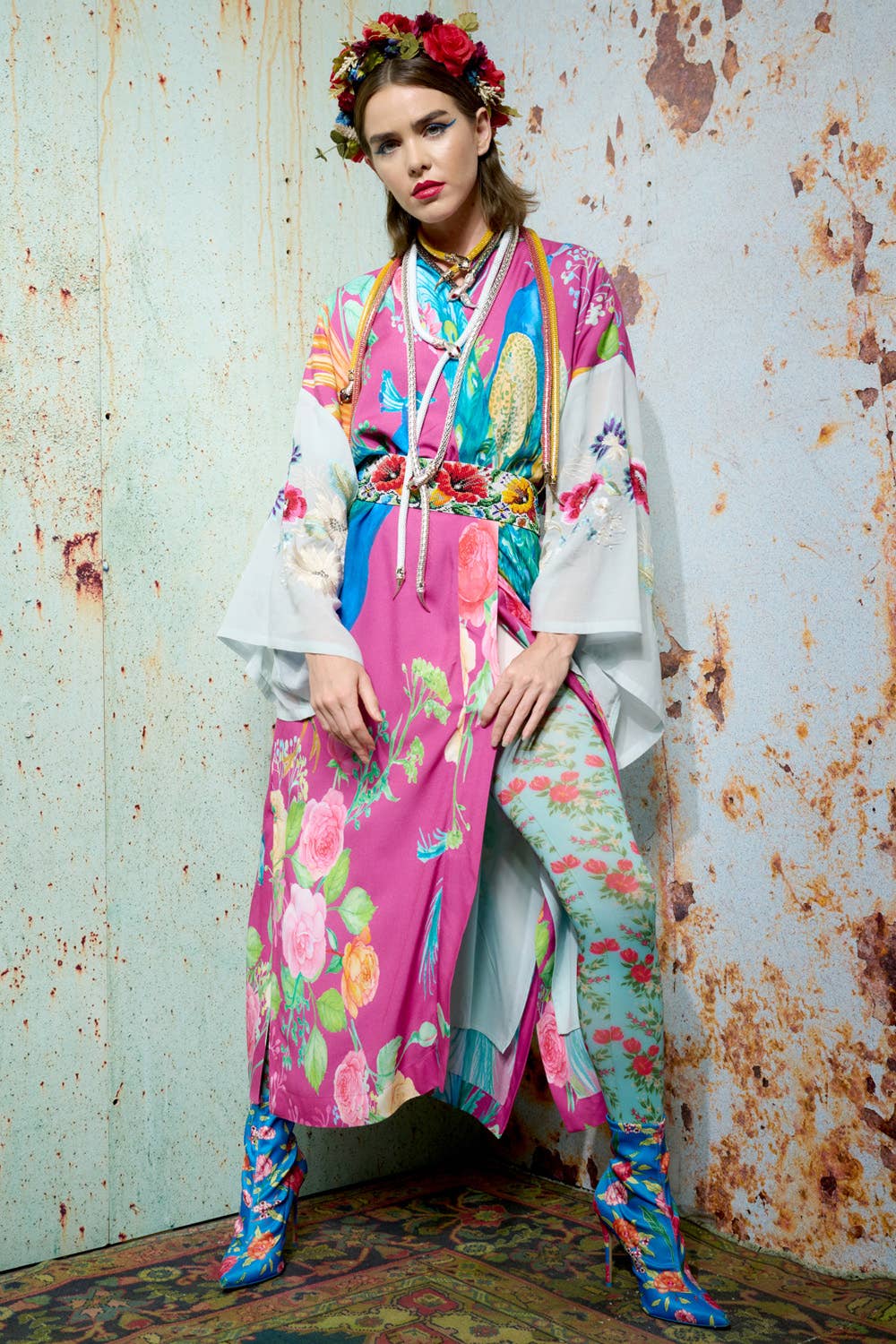 A woman stands against a textured wall wearing the vibrant Aratta Marie Antoinette Kimono with embroidered floral sleeves, a matching flower crown, blue floral boots, and boldly patterned flower motif tights in pink, blue, green, and yellow.
