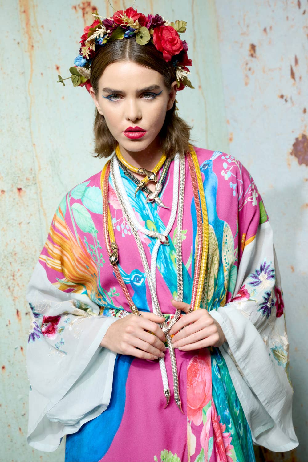 A woman in the Aratta Marie Antoinette Kimono with embroidered sleeves and a flower crown poses against a textured wall, gazing at the camera with bold makeup and decorative ropes around her neck.