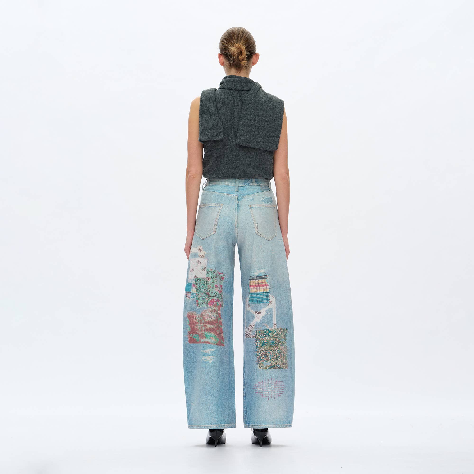 A woman stands facing away, wearing BAYEAS Light Blue Patch Pattern High-Rise Barrel Jeans with a sleeveless dark top, a matching sweater over her shoulders, black shoes, and her hair styled in a low bun.
