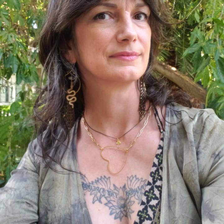 A woman with wavy brown hair poses outdoors, layered necklaces and Dreamers of Dreams’ Rising Snake Earrings accenting her look. A V-neck top reveals a floral and script chest tattoo, with green foliage in the background.