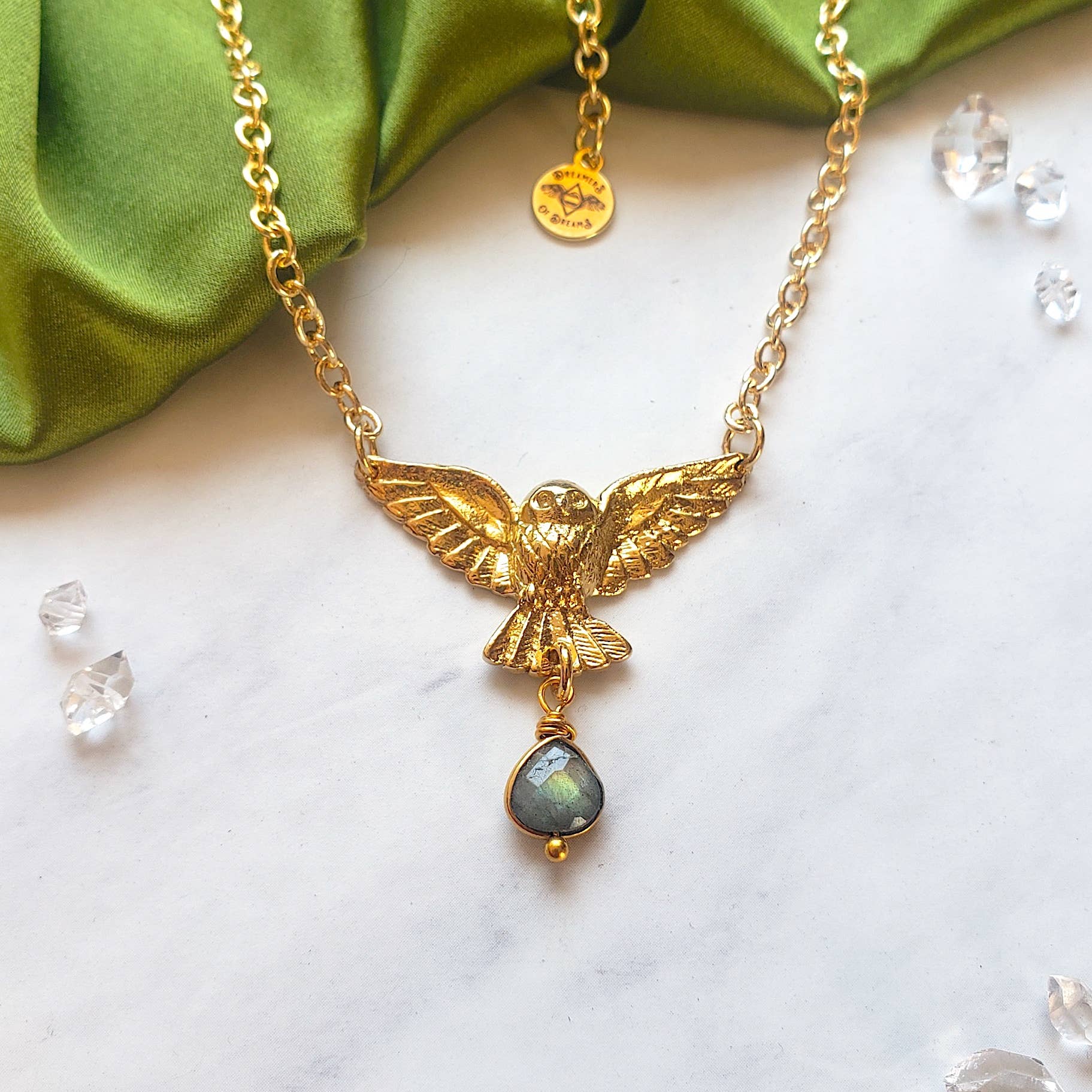 The Golden Owl of Intuition Labradorite Pendant Necklace by Dreamers of Dreams features a gold-plated owl with outstretched wings and a teardrop labradorite crystal, displayed on white with green fabric and crystal accents.