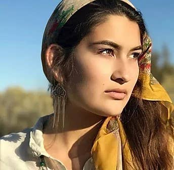 A woman with long brown hair wears a patterned headscarf, dangly earrings, and the Tara Gasparian Jewelry 32 Sacre Coeur Necklace, gazing thoughtfully to the side in sunlight against a blurred outdoor background.