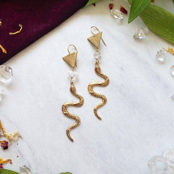 The Gold Year of the Snake Earrings by Dreamers of Dreams feature handcrafted snake designs with triangle tops and clear bead accents, showcased on white marble with scattered crystals, petals, and green leaves.