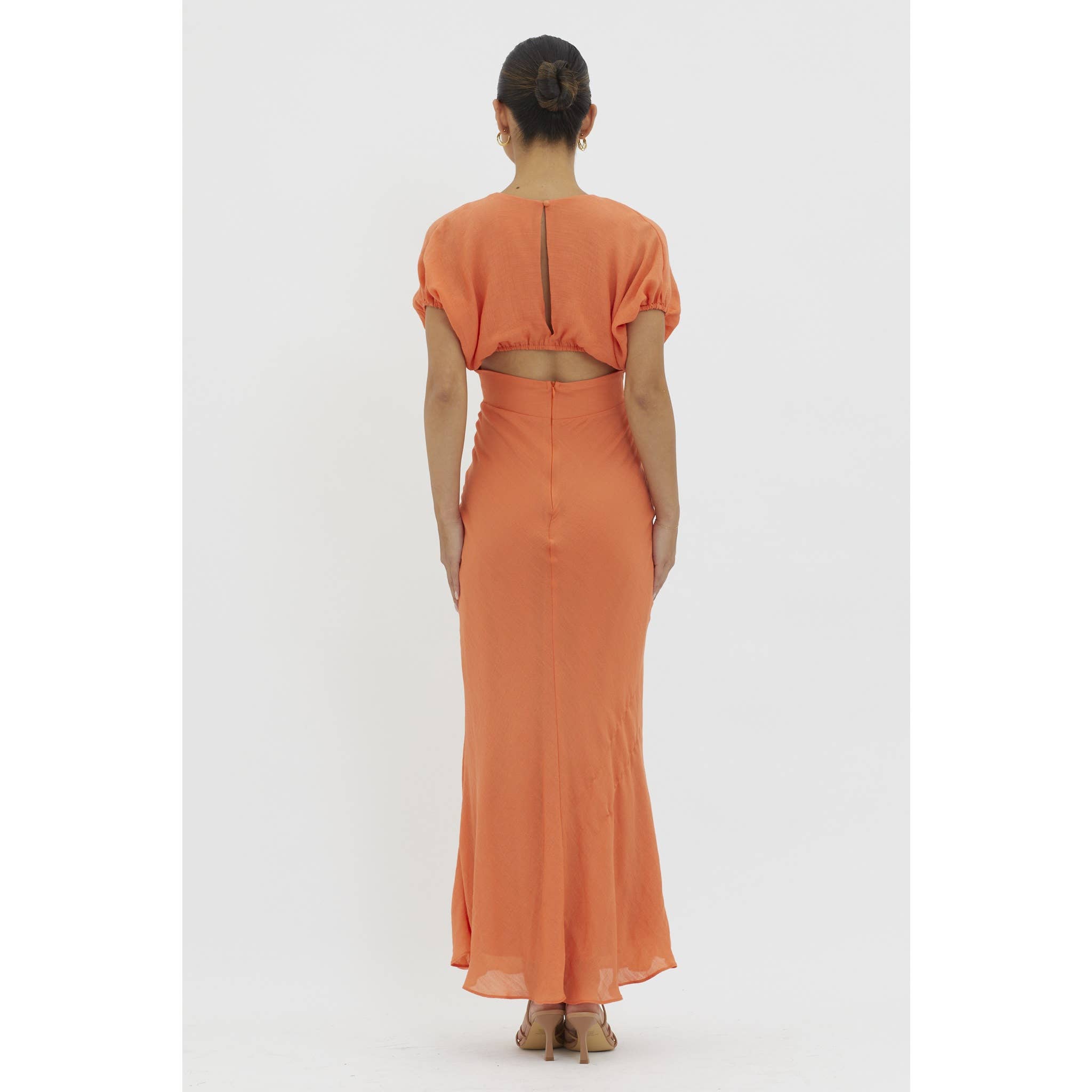 A woman with her hair in a bun stands facing away against a white background, wearing the ankle-length, short-sleeved Tangerine V-neck Maxi Dress with a keyhole back by One and Only Collective.