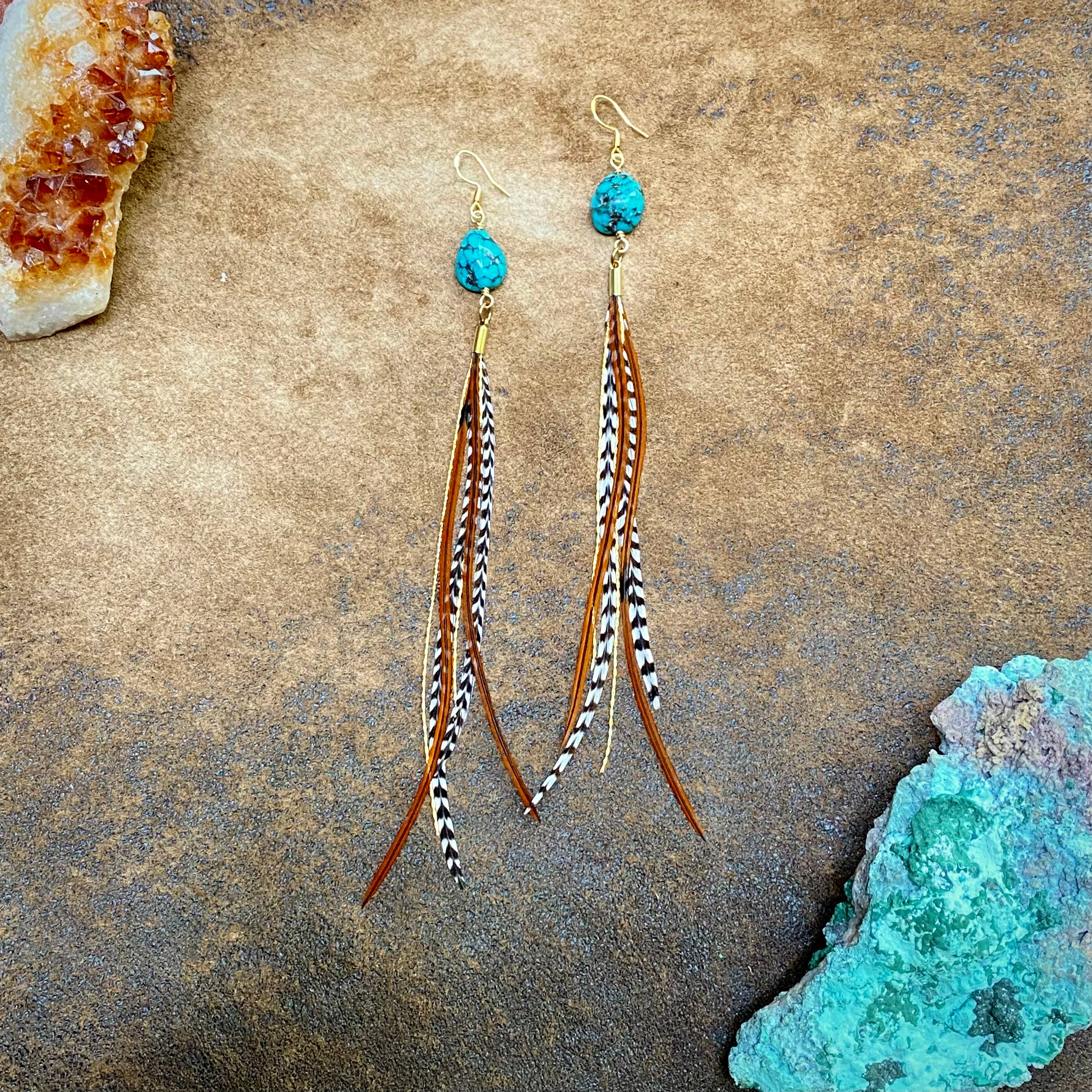 ASTALI Turquoise & Mini Feather Earrings - Grizzly & Fawn/Gold: Gold-plated earrings with turquoise beads and brown, black, and white feather-like strands, shown on a textured brown surface with mineral rocks.