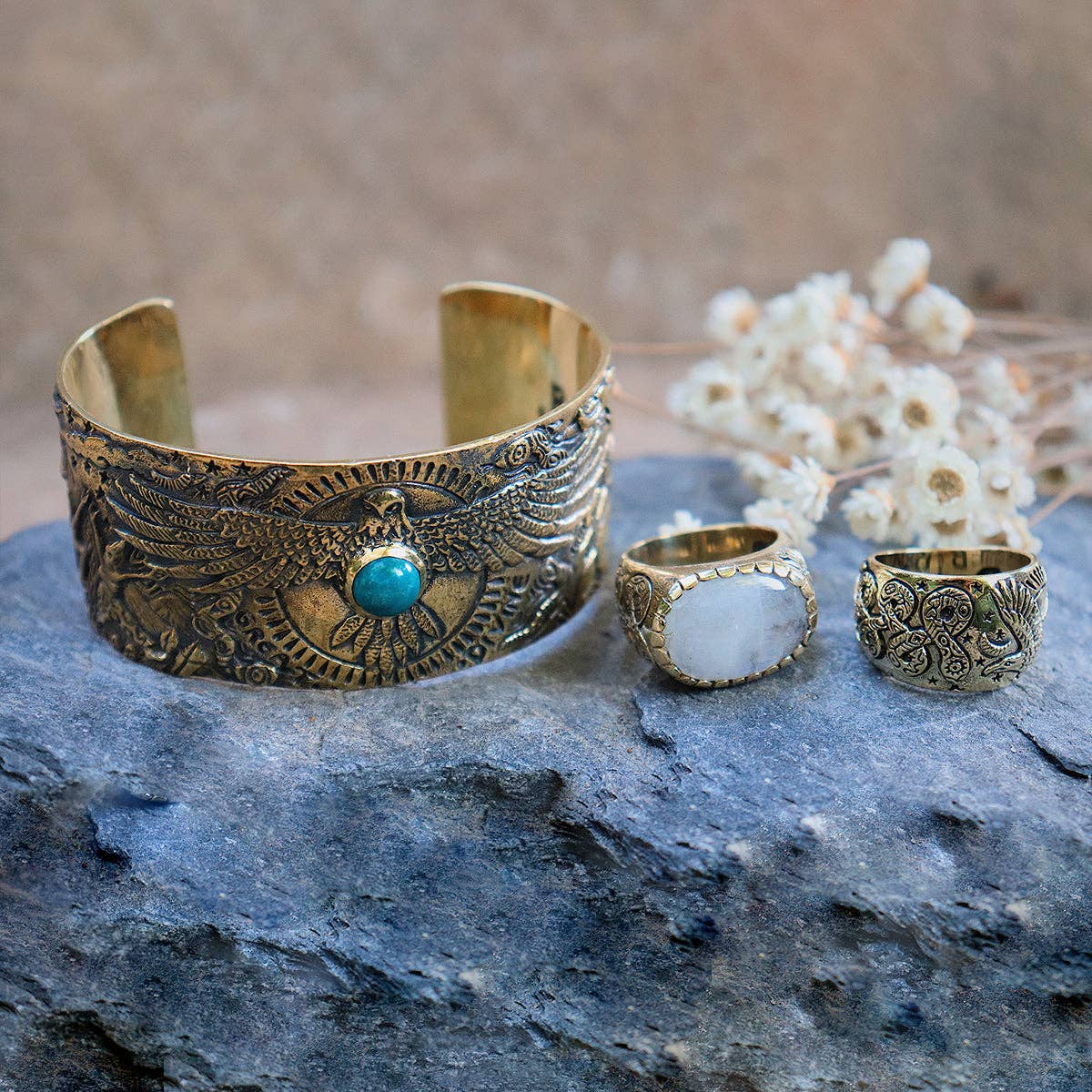The Rainbow Serpent Ring from bahgsu jewels, part of the Xavier Rudd Collab, is shown alongside an eagle gold cuff and a turquoise stone ring, all displayed on gray stone with dried flowers in the background.