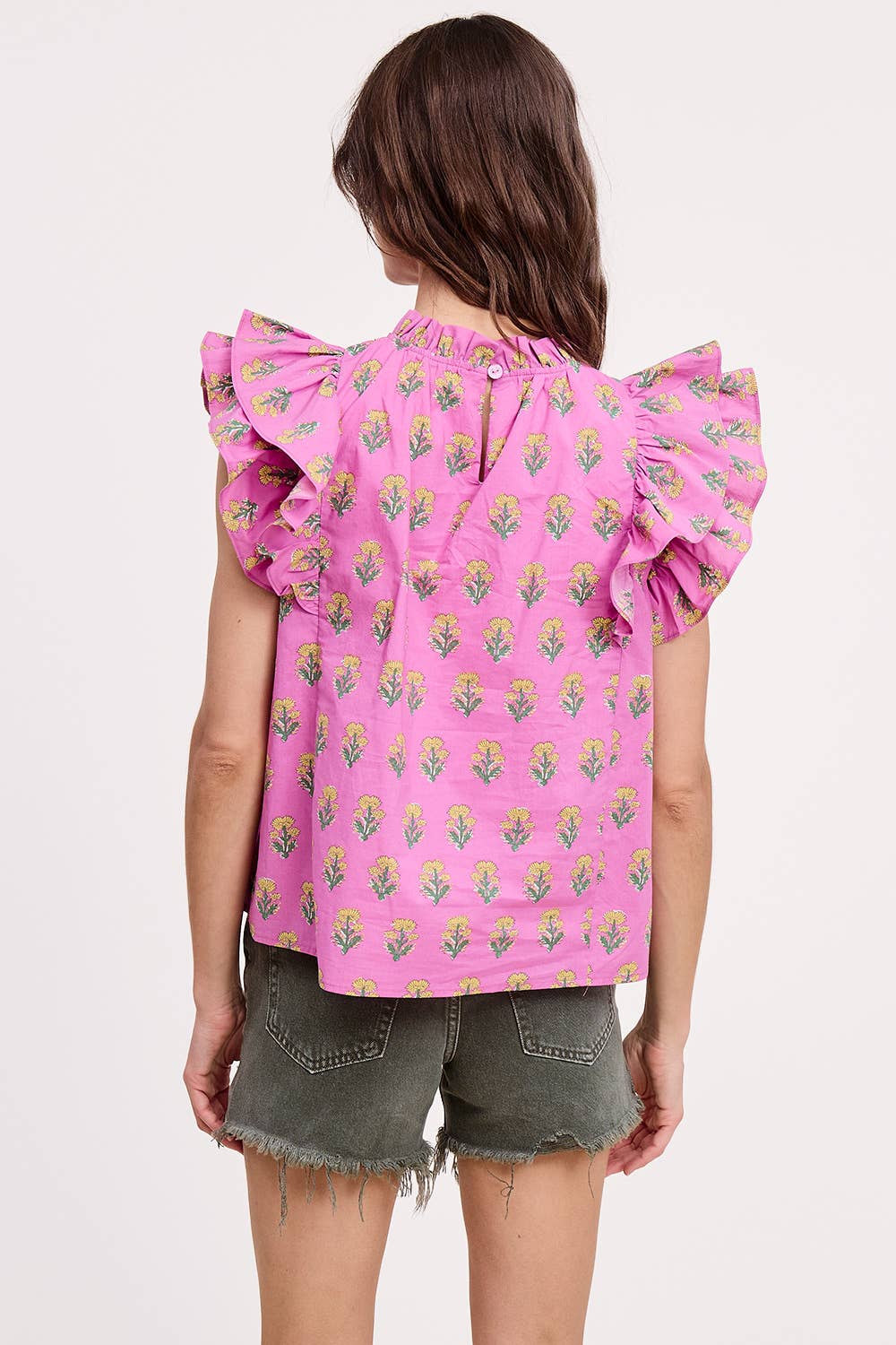 A woman with wavy brown hair is seen from the back wearing the Ultramarine Celeste Flutter Sleeve Blouse in pink floral print and frayed green denim shorts, set against a plain white background.