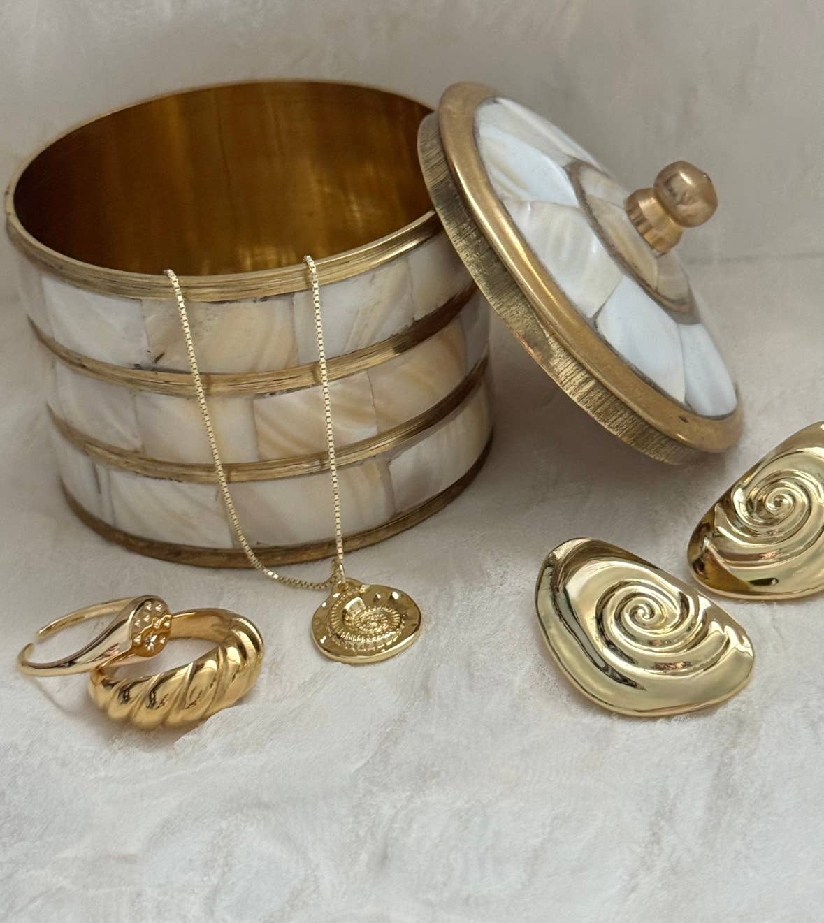 A gold and white decorative jewelry box with its lid off displays the Mar Medallion Necklace by Tramps+Thieves, along with a chunky gold ring and matching gold swirl earrings on a light textured surface.